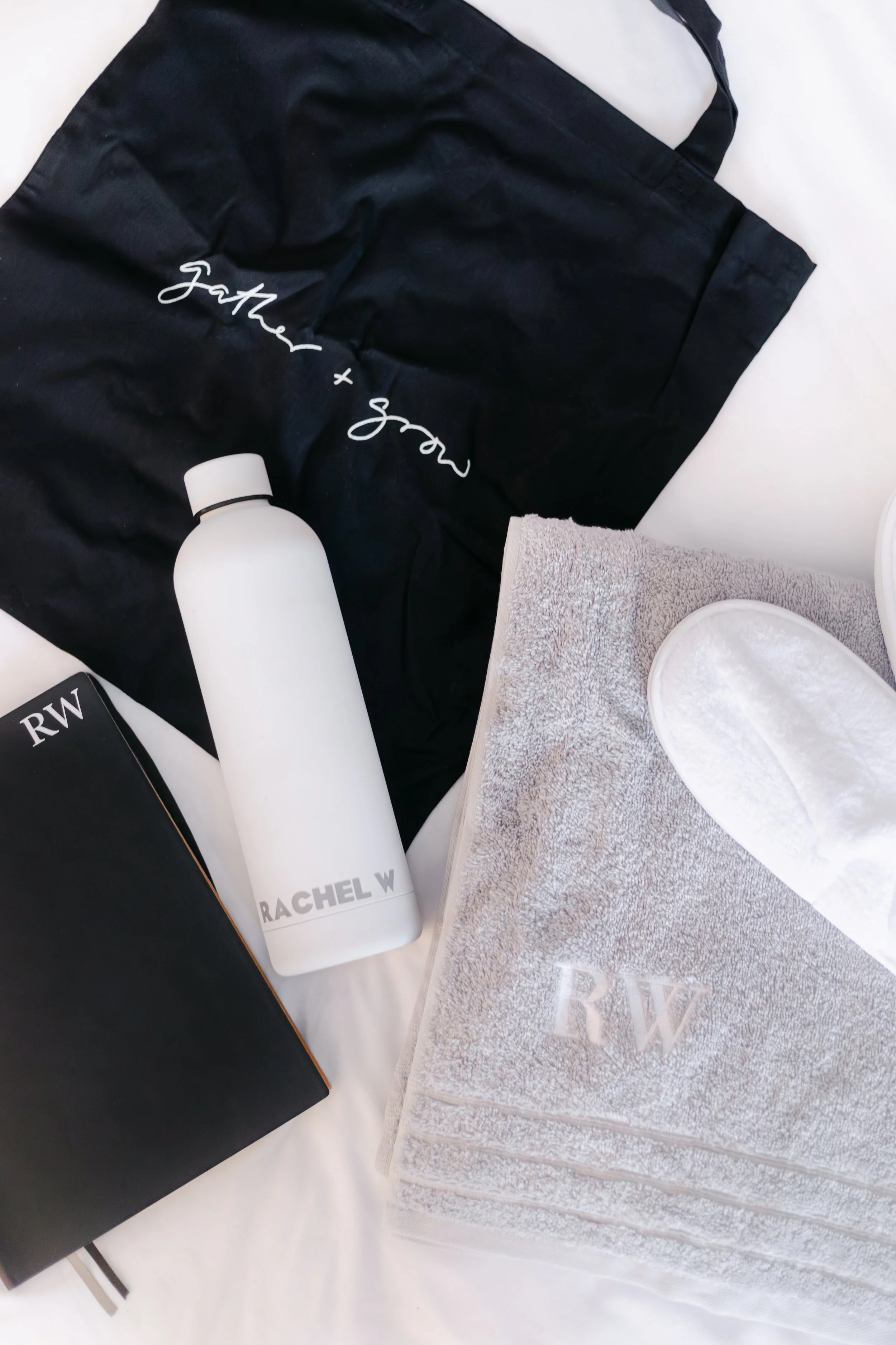 A collection of luxury personal care and clothing items, including a black tote bag with white script, a white water bottle with gray text, a gray towel with embroidered initials, a black box, and a white facial scrubber, all arranged on a white surf