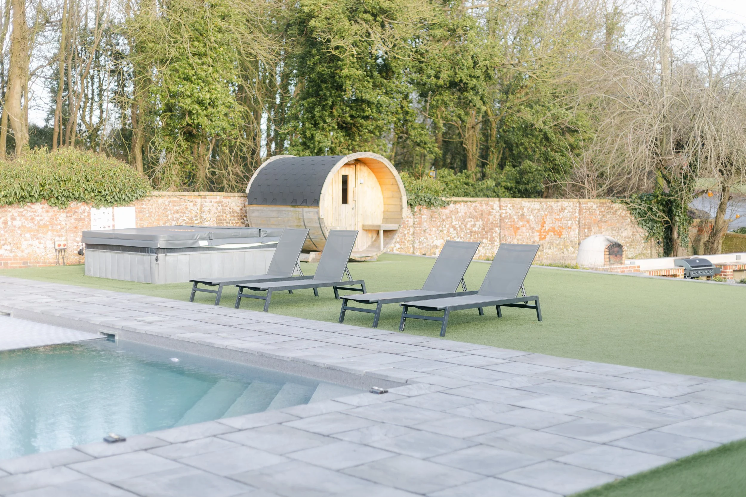 Outdoor backyard with a swimming pool, four lounge chairs, a hot tub, a wooden barrel sauna, a brick oven, and trees in the background.