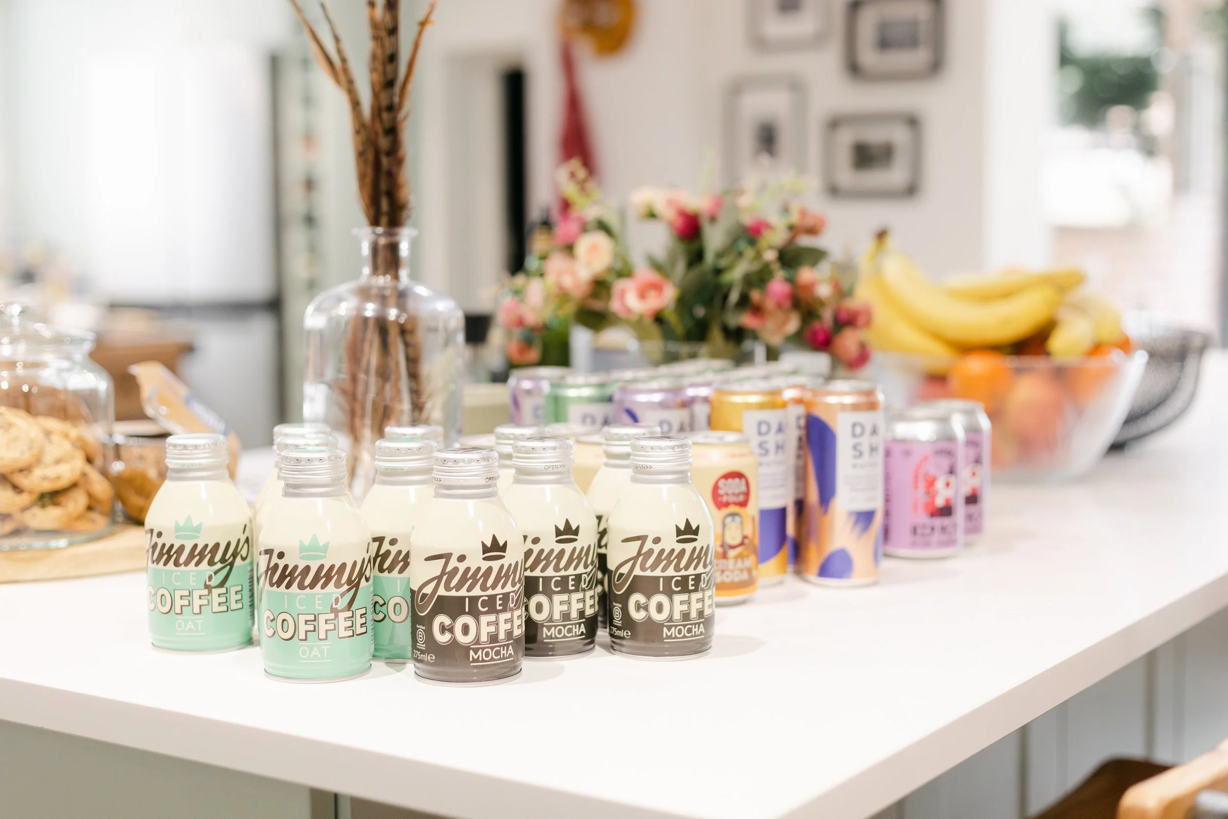 Assorted canned and bottled drinks, including Jimmy's Ice Coffee, and a bowl of bananas and other fruit on a white kitchen countertop. In the background, there is a vase with flowers and a small jar of cookies.