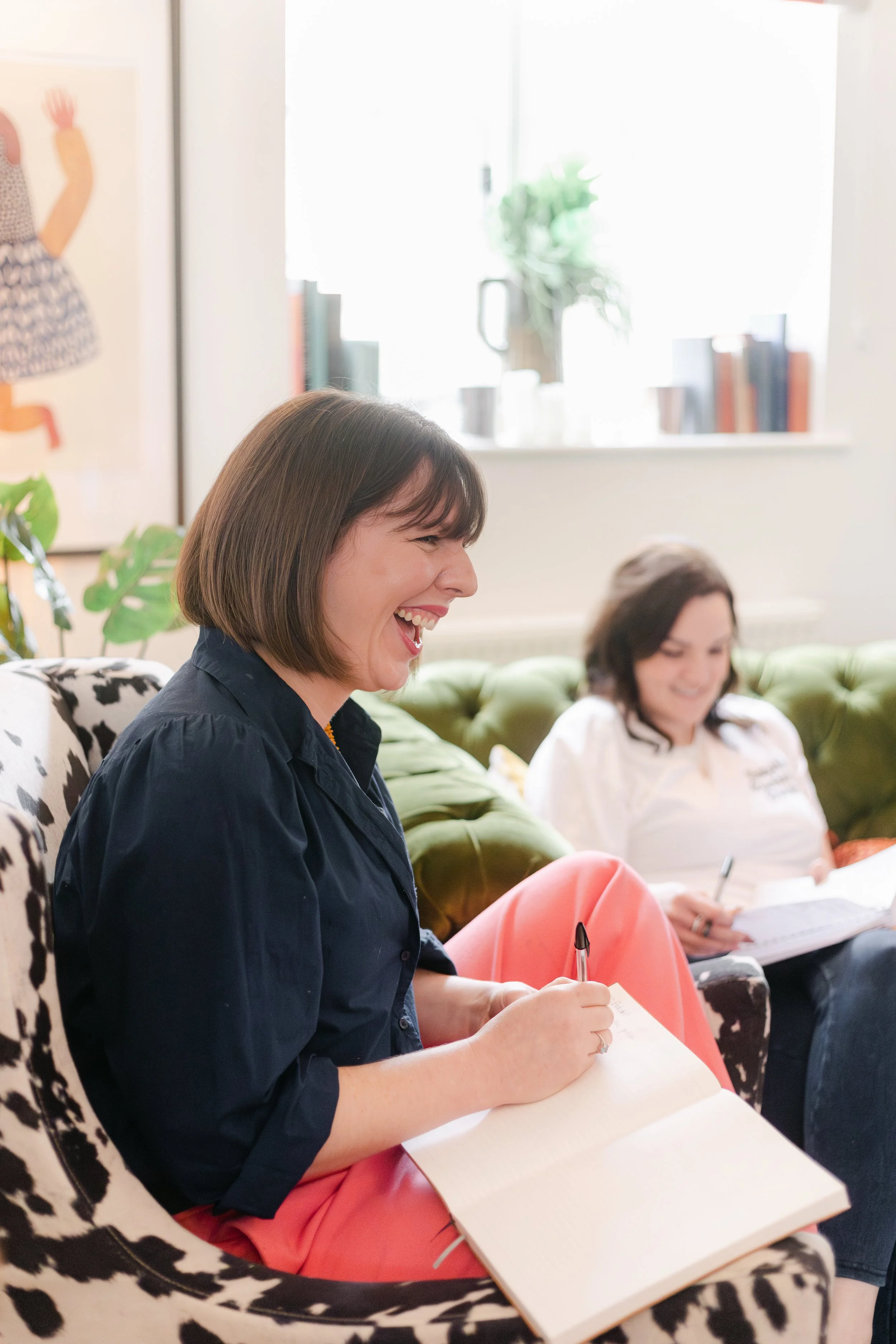 Two women sitting on a patterned and green couch in a bright room, laughing and taking notes during a meeting or discussion.