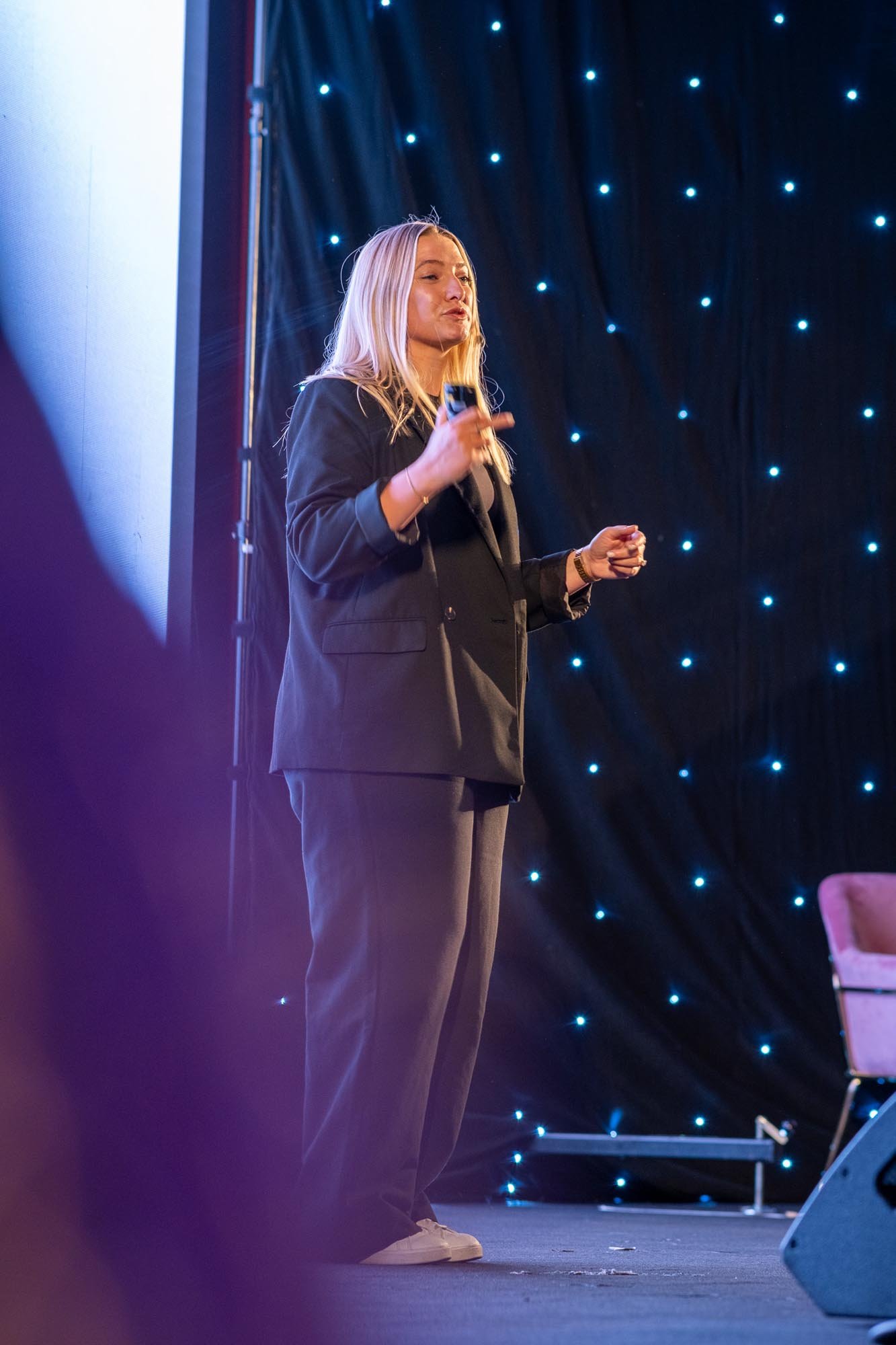A woman with blonde hair in a black blazer and gray pants standing on a stage with a black background decorated with blue lights, holding a microphone.