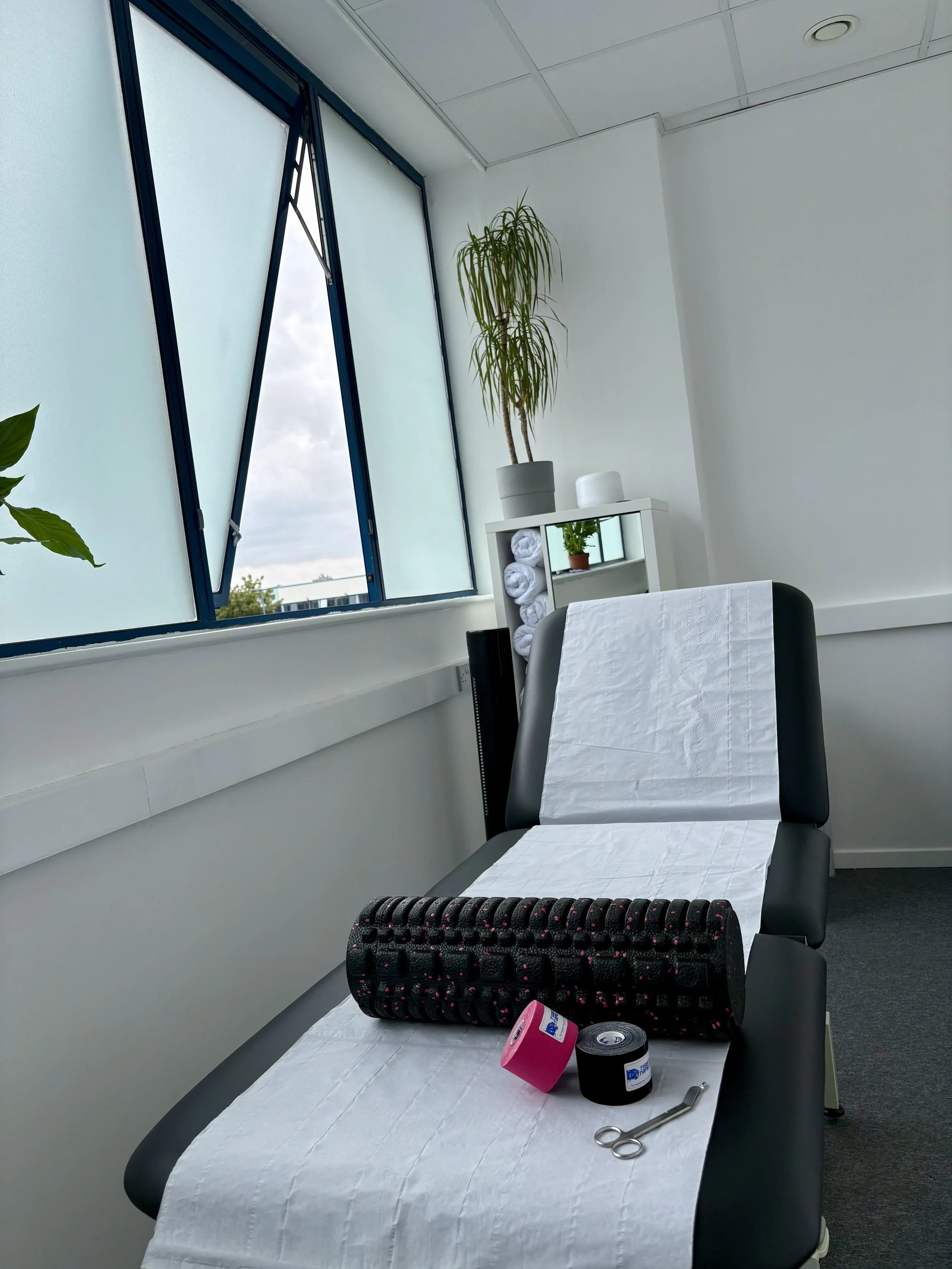 A clinic room with a black examination bed covered with a white paper sheet. On the bed are a foam roller, pink and black kinesiology tapes and a small pair of scissors. In the background, there is a white shelf with rolled towels and a potted plant.