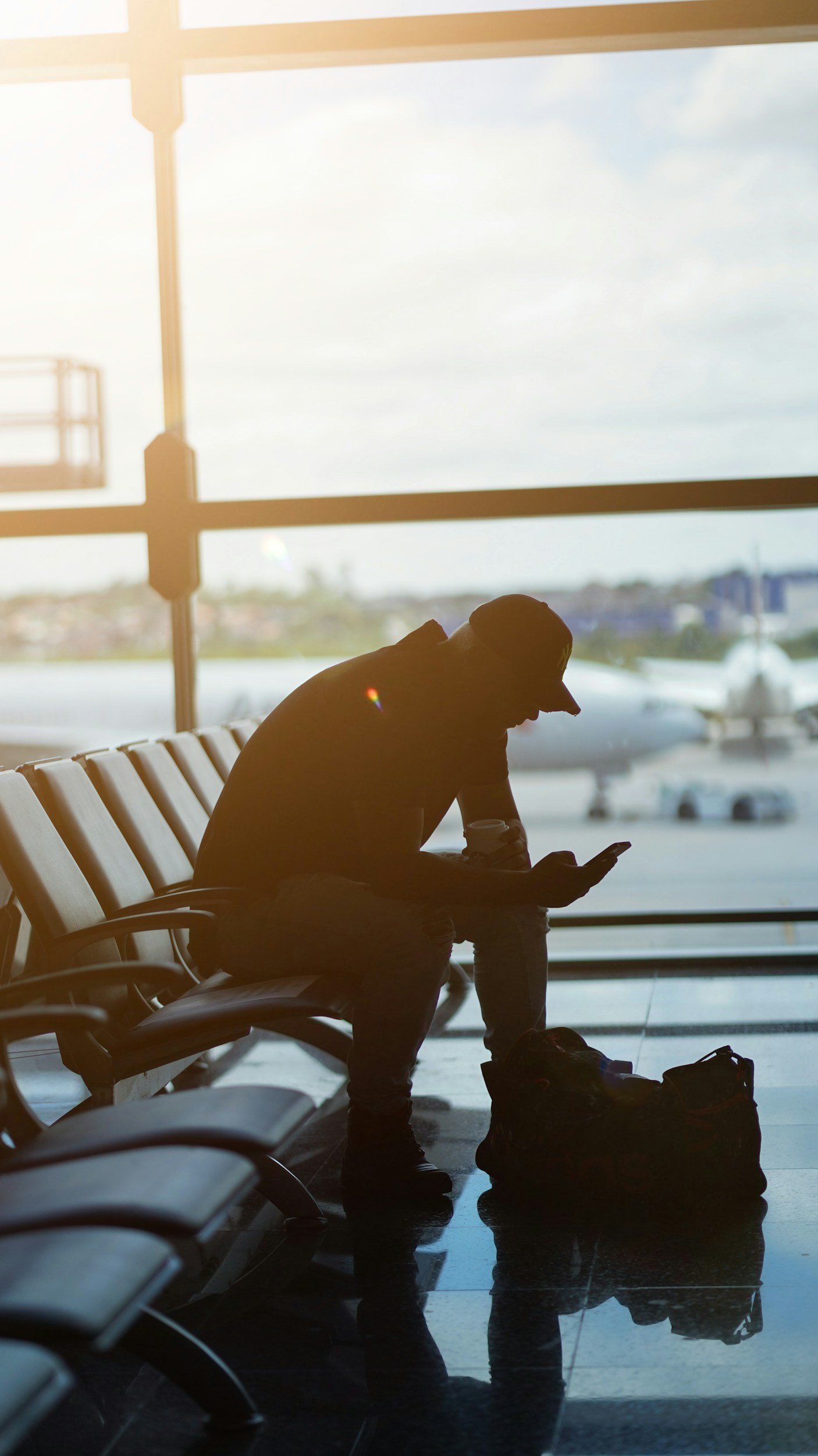 Silhouette of a person in an airport waiting area, sitting on a bench, looking at their phone, with luggage on the floor in front of them, and planes visible outside the large window.