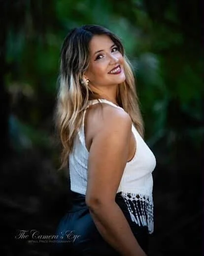 A woman with long, wavy hair wearing a white sleeveless top and smiling, standing outdoors with blurred green foliage in the background.