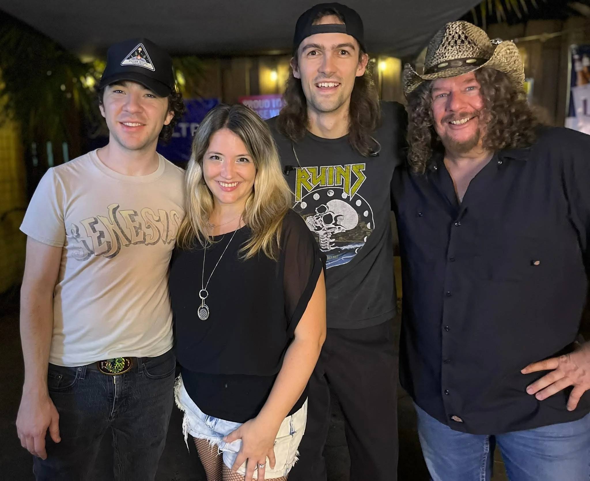 Five people standing together and smiling at a social event, with two men wearing hats, one woman with blonde hair, and two men with long hair, one wearing a bandana.