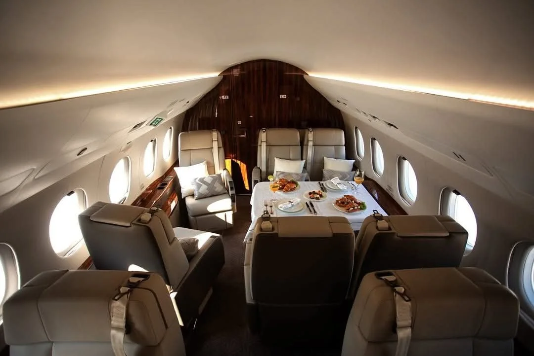 Inside a private airplane cabin, with beige leather seats, a dining table set with plates, glasses, and food, surrounded by small oval windows, and warm lighting.