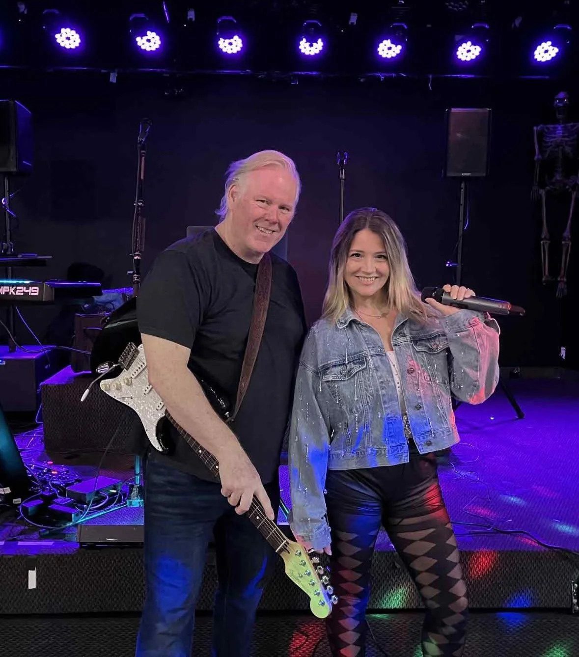 A man and woman on a stage with musical equipment, smiling for the camera. The man holds a guitar, and the woman holds a microphone. The background is lit with purple stage lights.