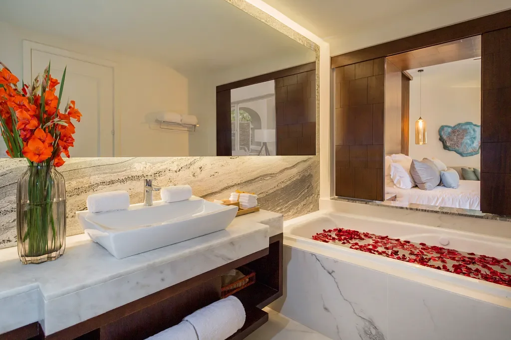 Bathroom with a marble vanity, vase with orange flowers, a bathtub filled with water and rose petals, and a large mirror reflecting part of a bedroom.