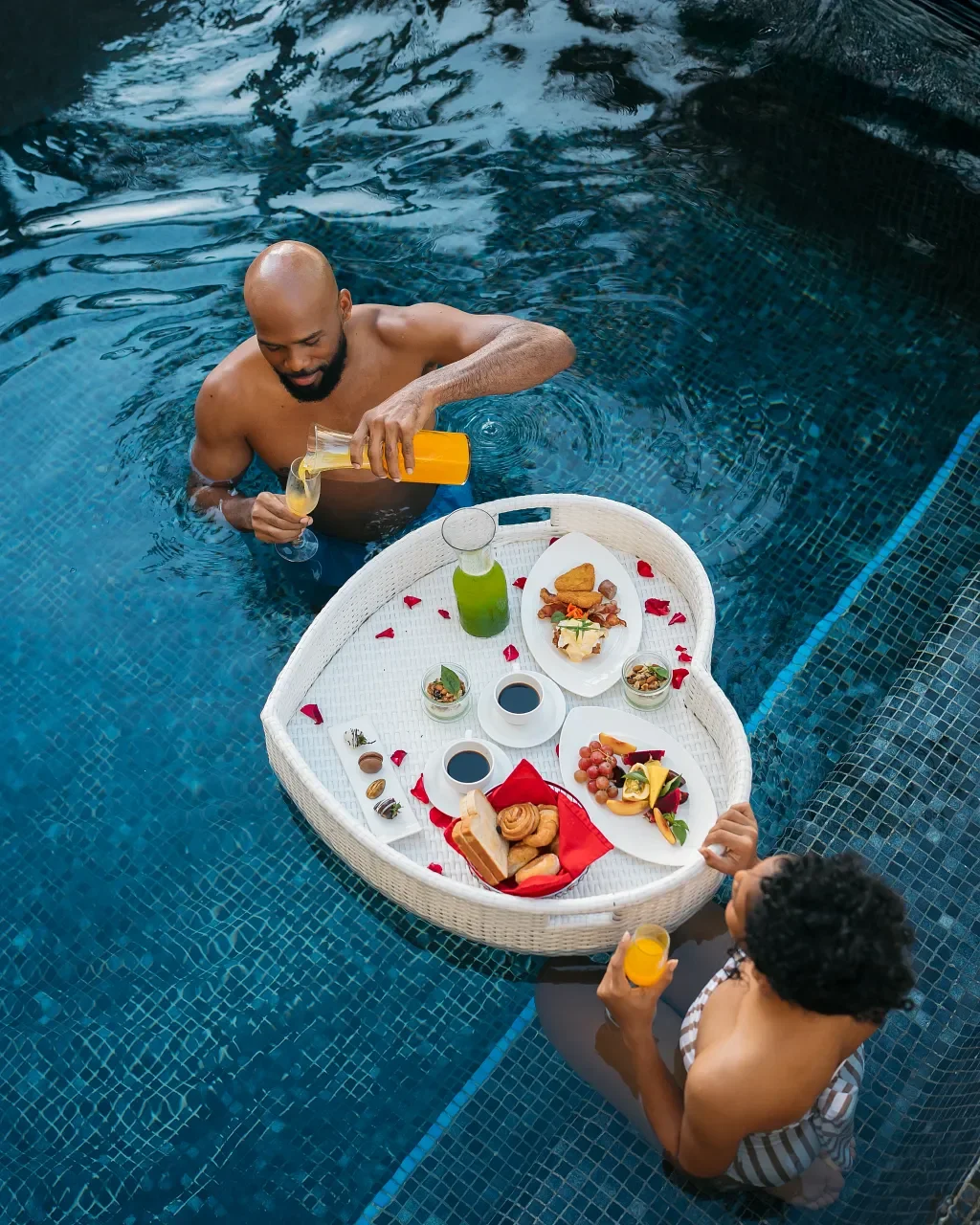 A man and a woman enjoying a floating breakfast with drinks in a swimming pool. The tray holds a variety of foods, drinks, and desserts.