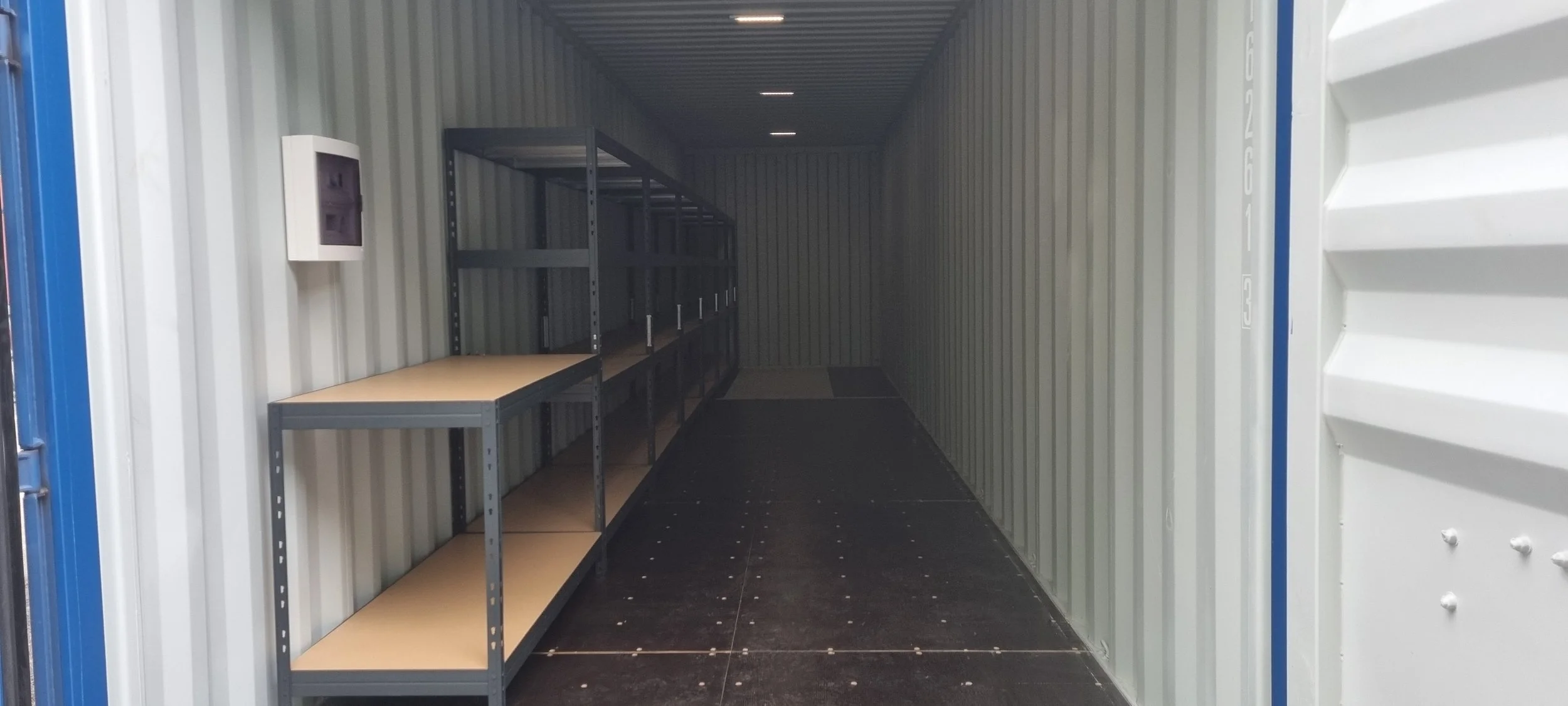 Empty storage unit with metal shelving and wood shelves on the left side, black floor, metal walls, and a small white file cabinet on the wall.