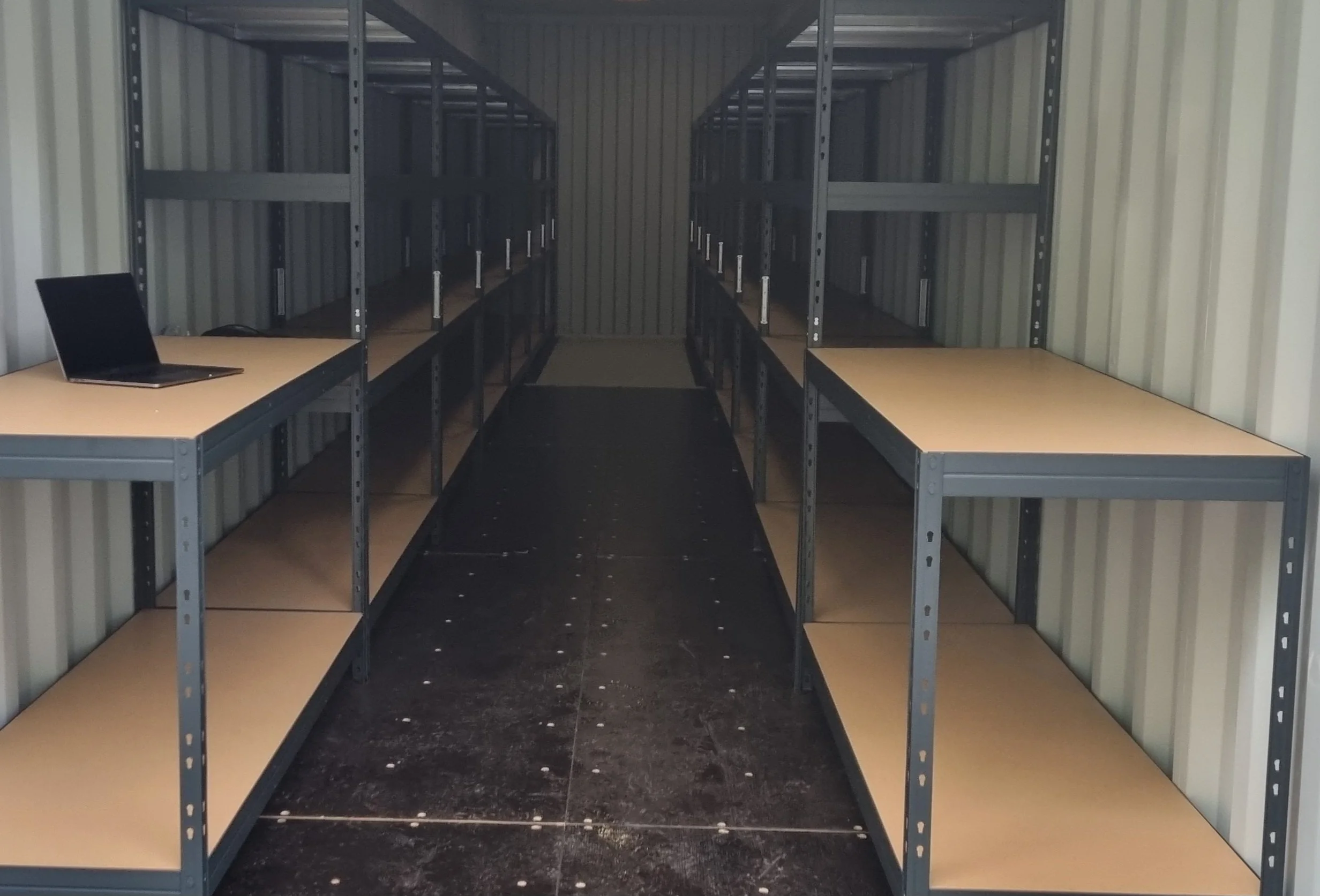 Empty storage container with metal shelves and two desks, one with a laptop, arranged along the sides.