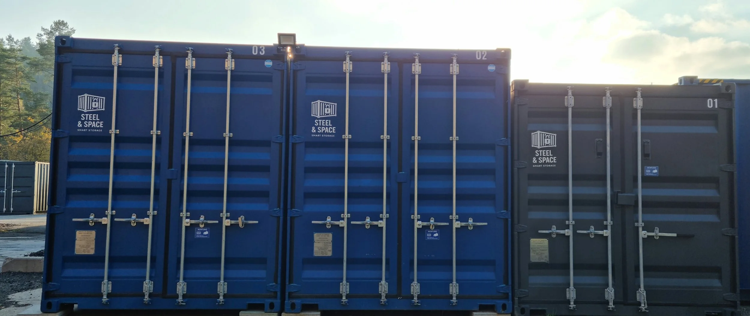 Two large shipping containers, painted blue and gray, with 'Steel & Space' logos, stacked side by side outdoors with trees and sky in the background.