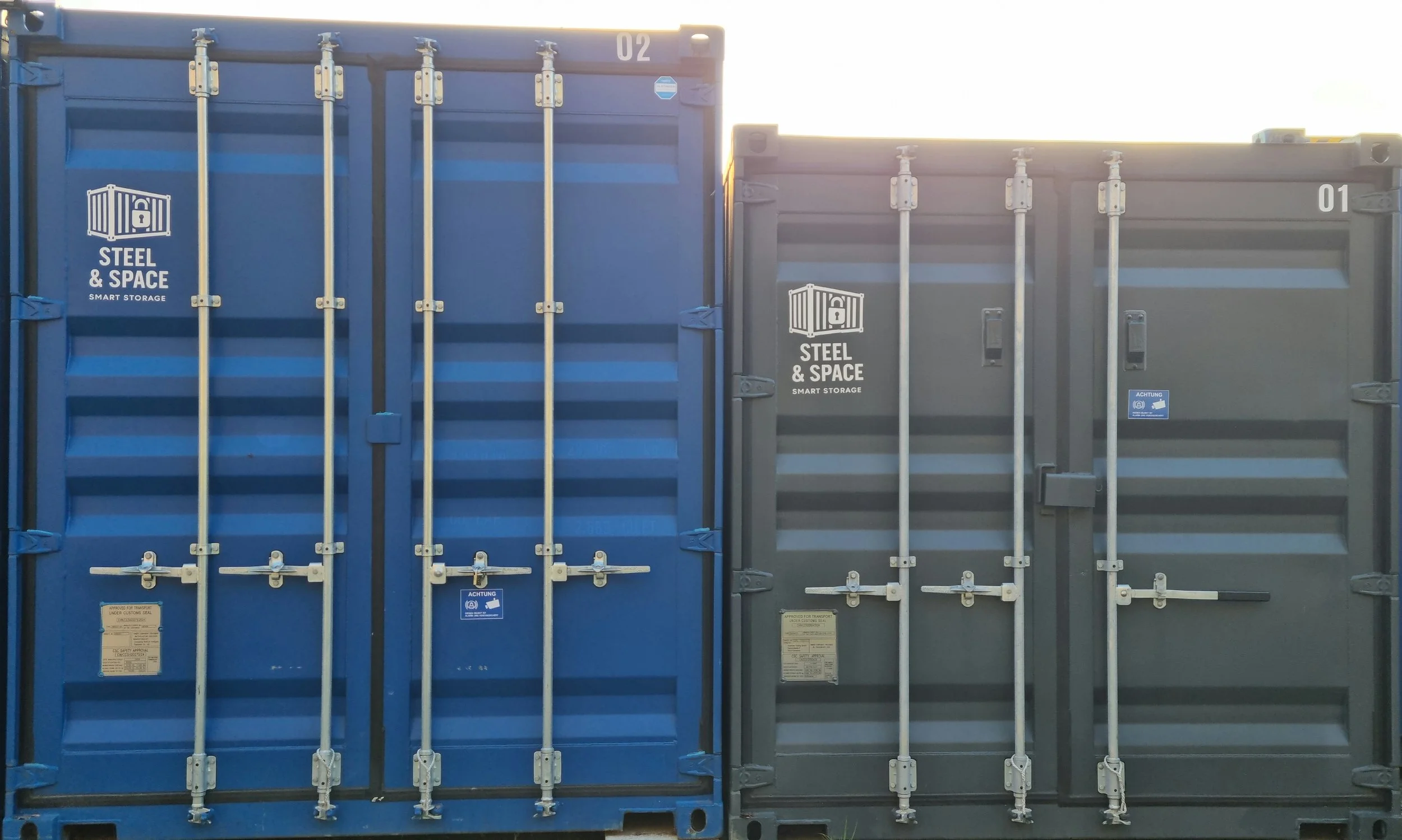 Two shipping containers, one blue and one gray, with the words 'Steel & Space Smart Storage' on their doors. The containers are stacked closely together with vertical locking bars.