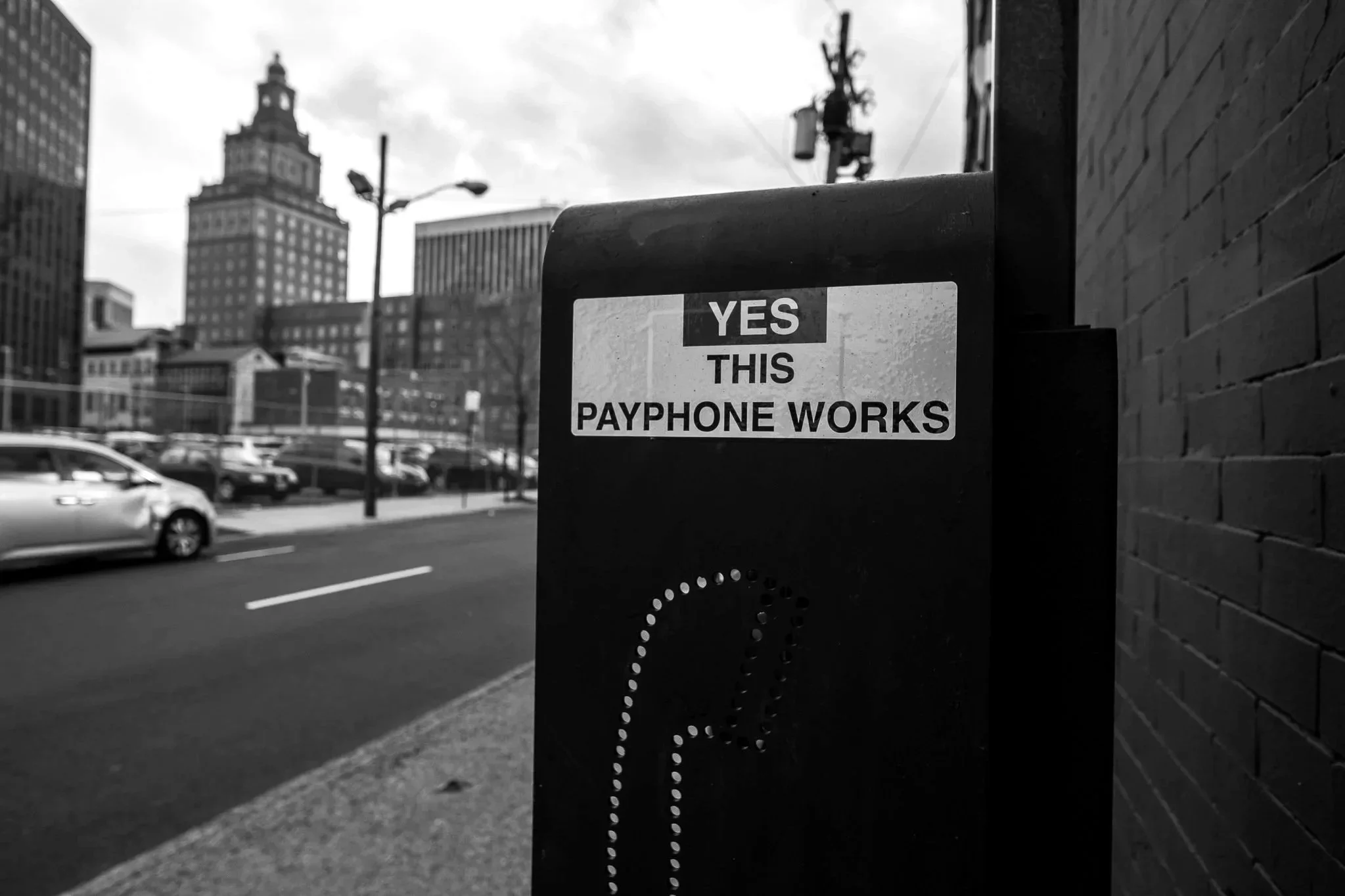 A city street scene with a payphone booth on the sidewalk, featuring a sign that says 'YES THIS PAYPHONE WORKS'. Parked cars are on the street, and tall buildings and utility poles are in the background under a cloudy sky.