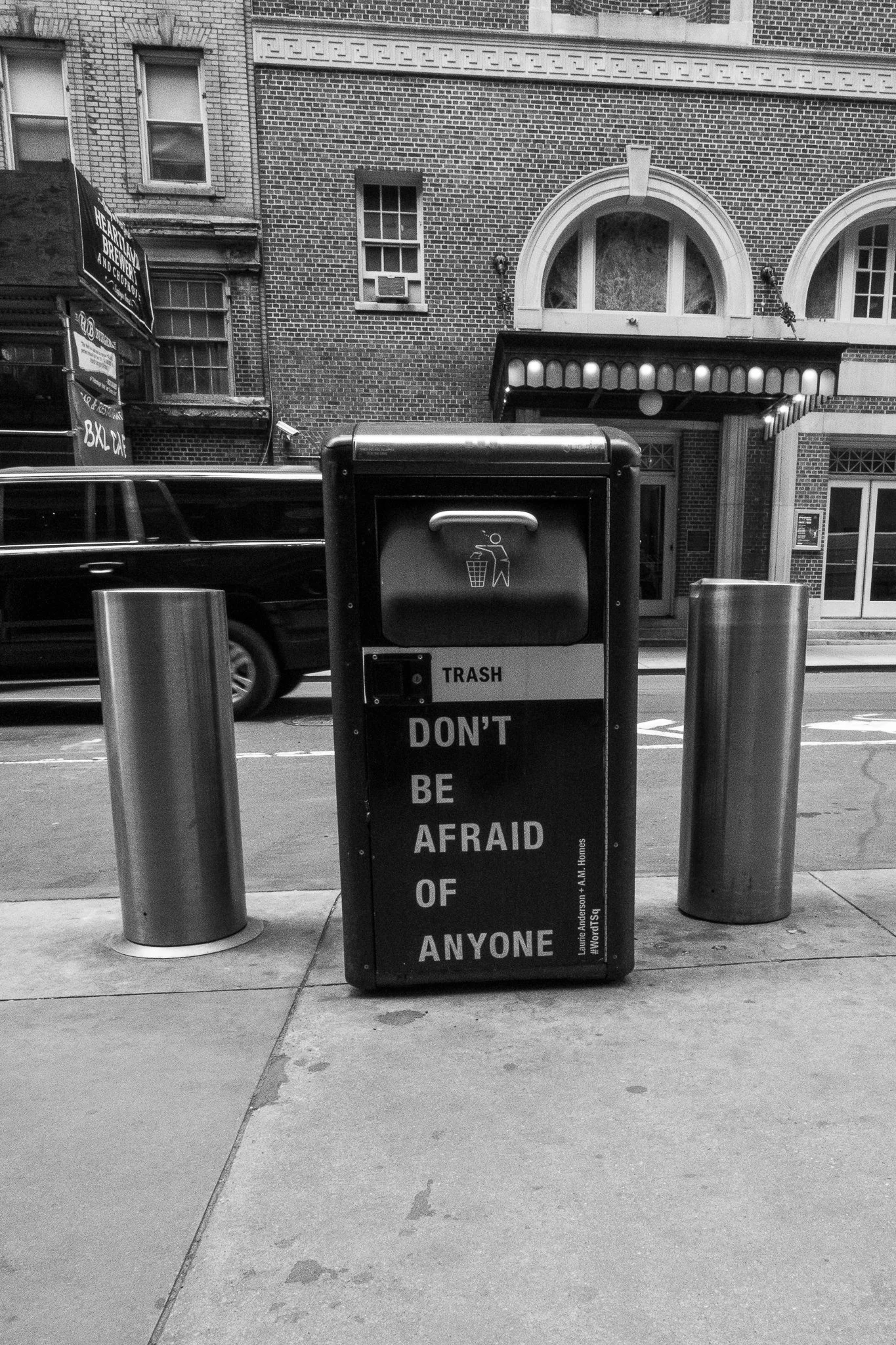 A city sidewalk scene in black and white, with a trash bin that reads 'DON'T BE AFRAID OF ANYONE' in the foreground, and a brick building with arched windows and a canopy behind it. A car is passing by in the background.