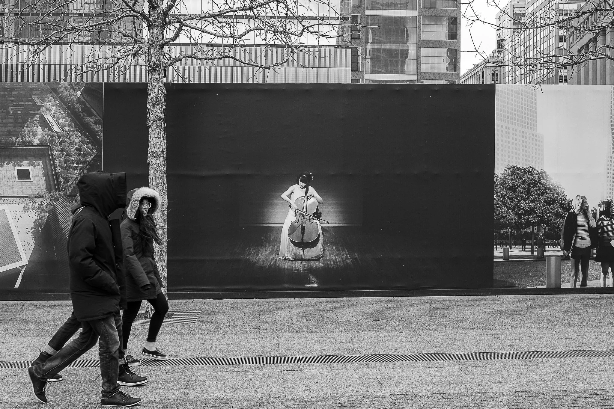People walking past a large outdoor poster featuring a female cellist performing in an urban setting, with high-rise buildings and leafless trees in the background.
