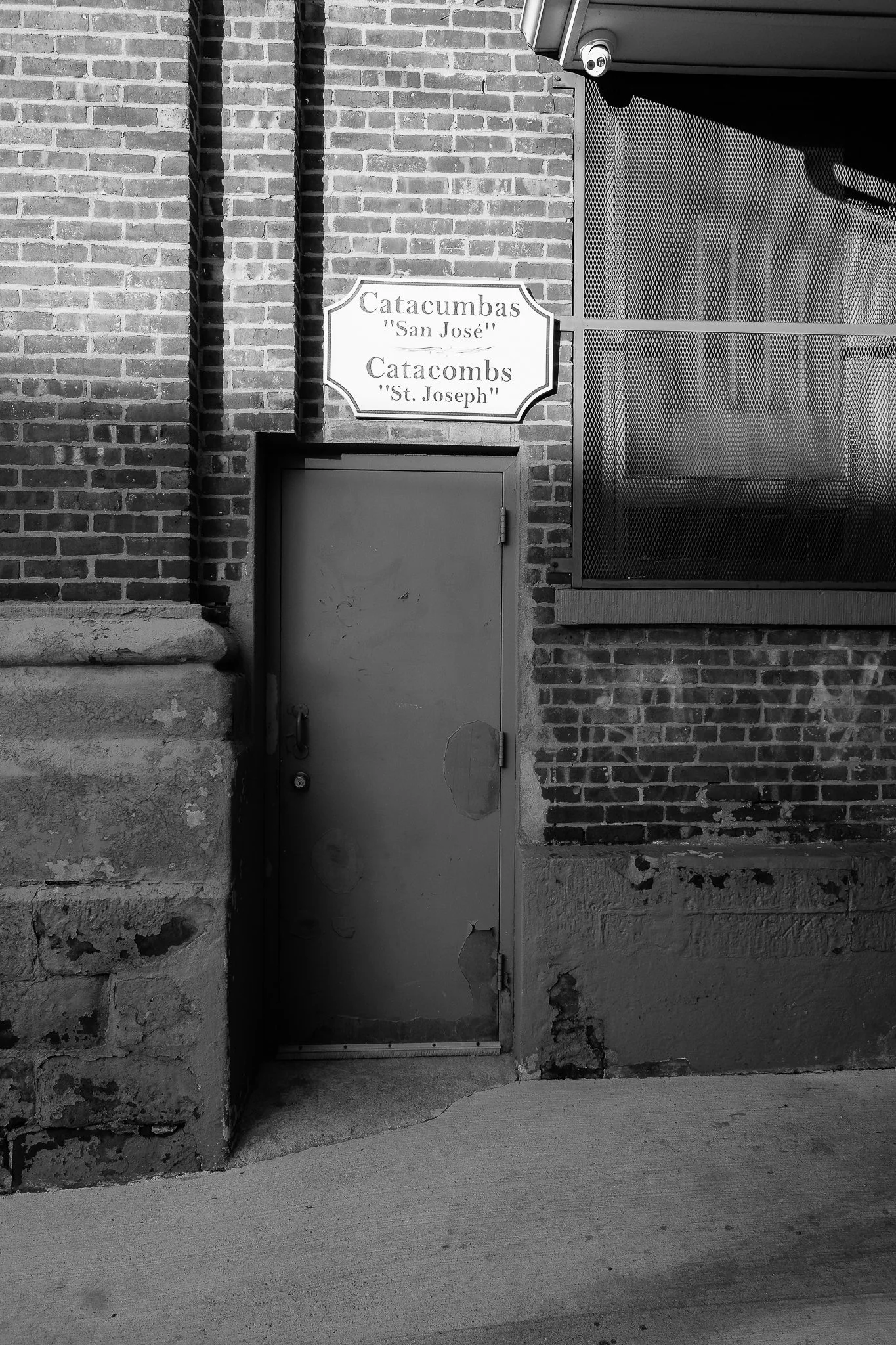 Black and white photo of a brick wall with a door, a sign above the door reads 'Catacumbas St. Joseph', and a surveillance camera is mounted near the top right corner.