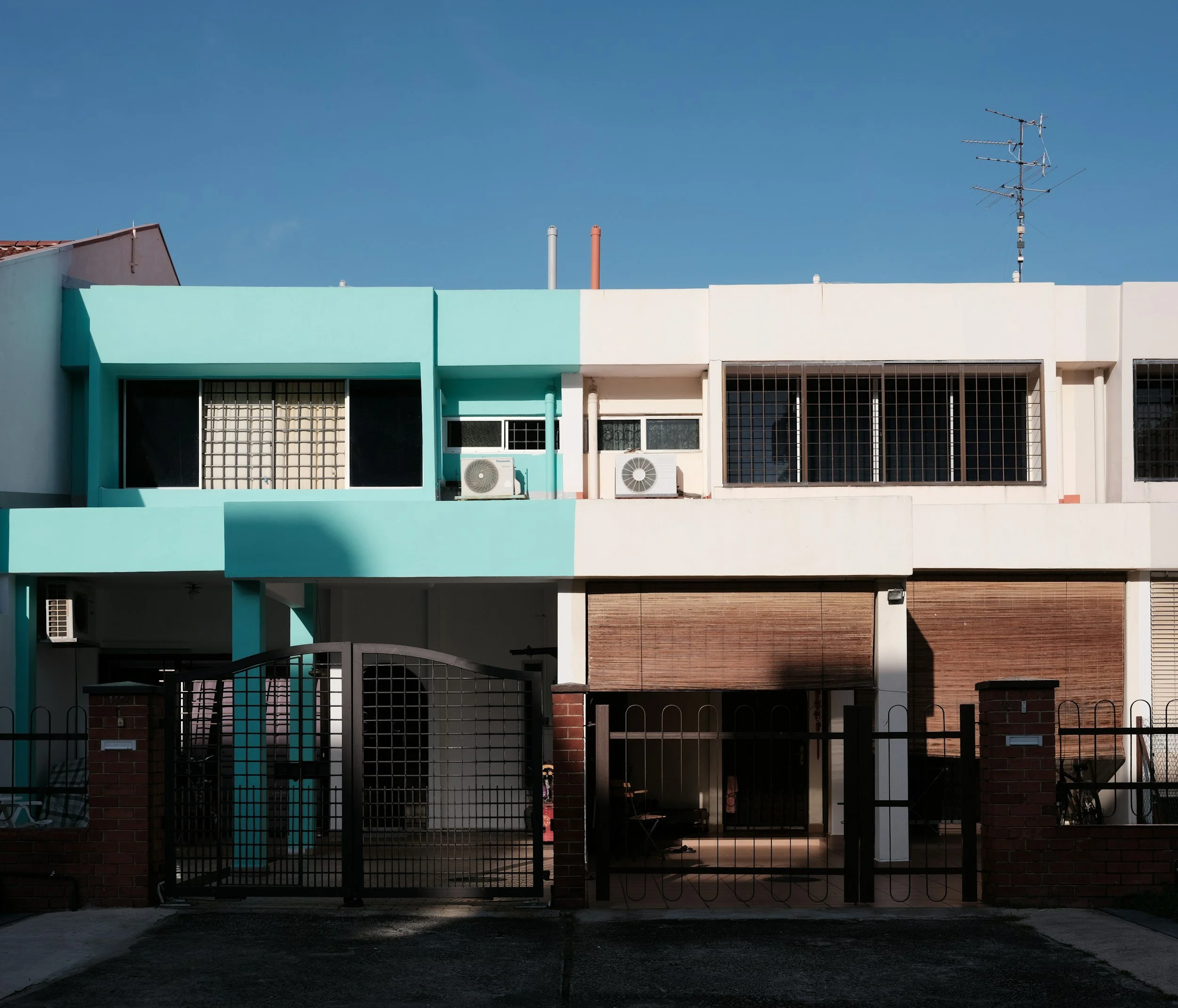 Front view of a two-story landed private residential building with a black gate, brick pillars, turquoise and white walls, and window air conditioning units. Clear blue sky in the background.