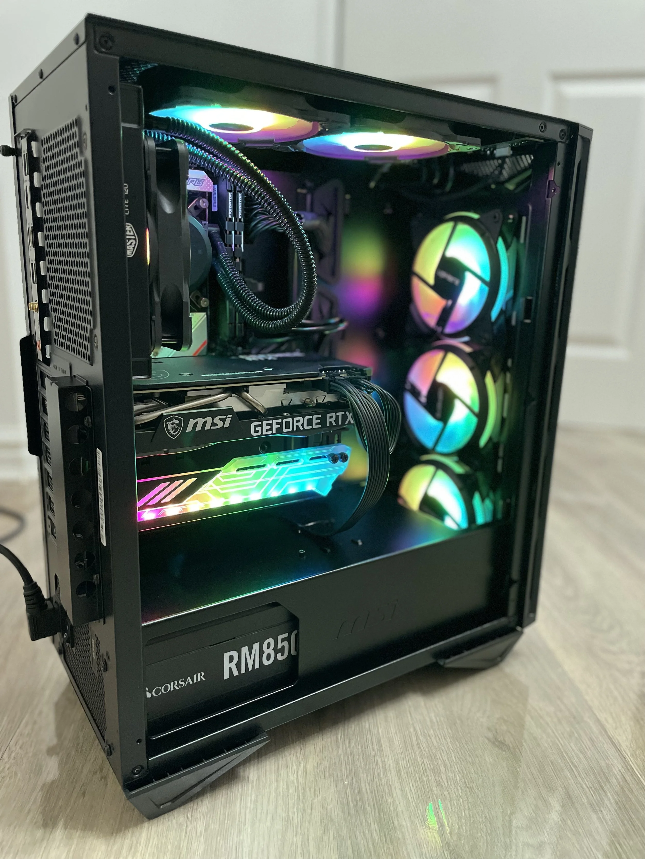 A gaming computer with a transparent side panel showing internal components, including a CPU cooler with RGB lighting, multiple RGB case fans, a MSI GeForce RTX graphics card, and a Corsair RM850 power supply, all illuminated with rainbow-colored RGB