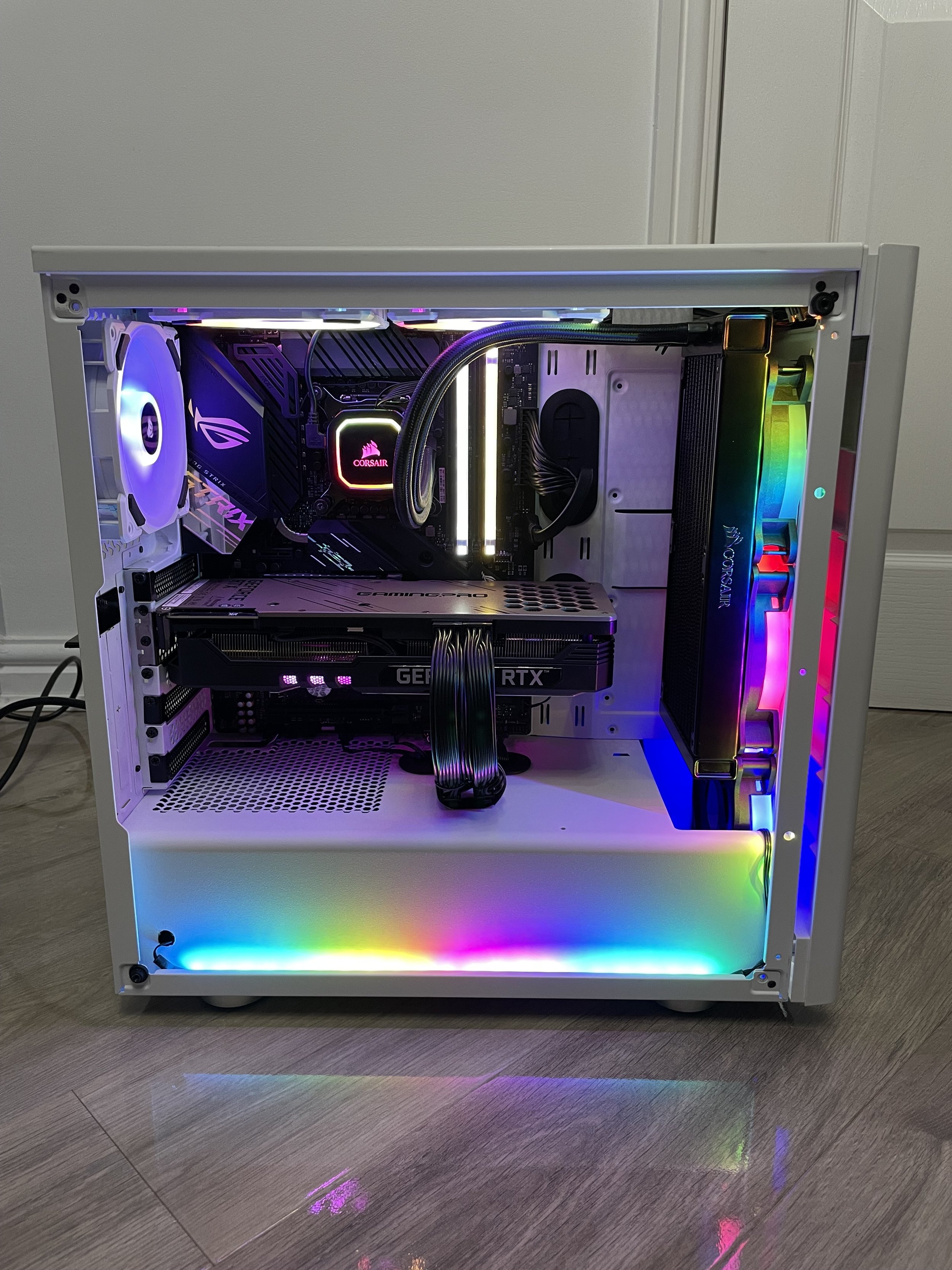A custom gaming PC with rainbow-colored RGB lighting, a liquid cooling system, a white case, and a graphics card labeled 'GEFORCE RTX'.