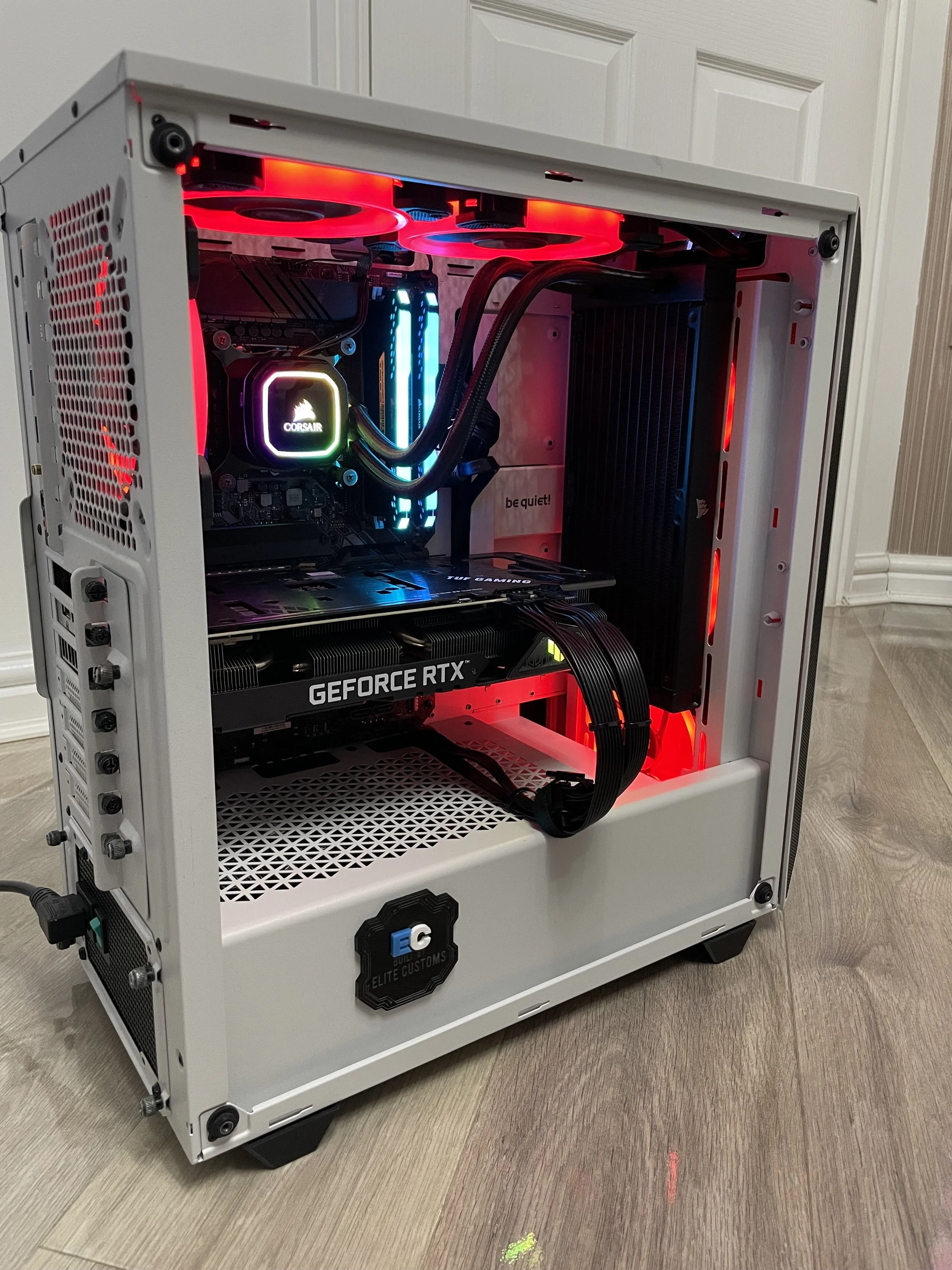 White gaming PC case with visible components including a Corsair cooler, RGB RAM, and a GeForce RTX graphics card, with red and blue internal LED lighting.