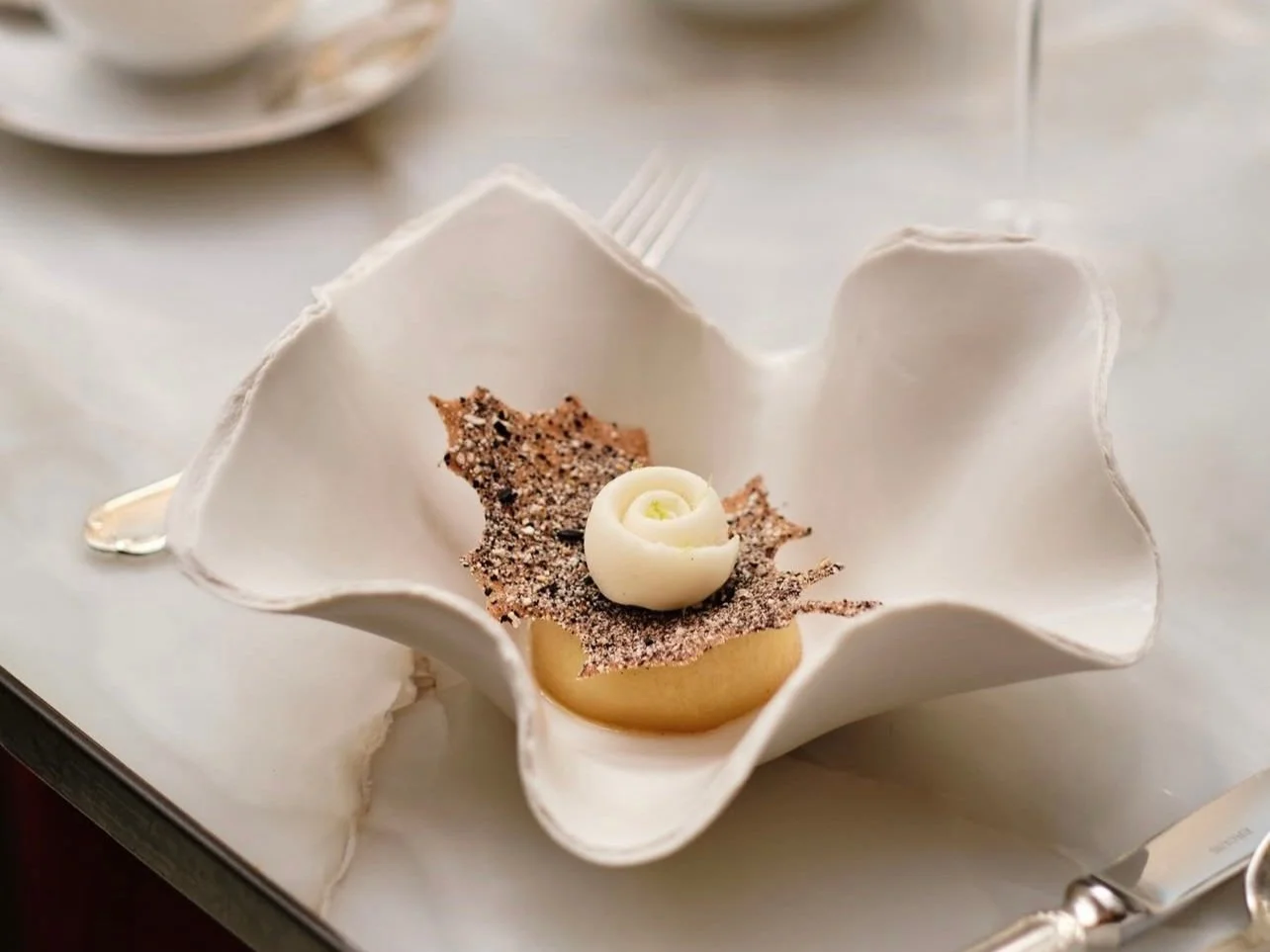 Dessert présenté dans une coupe en forme de fleur blanche contenant une mousse ou un gâteau accompagné de décoration en chocolat et d'une petite rose blanche en chocolat ou en pâte de sucre, sur une table avec des couverts.