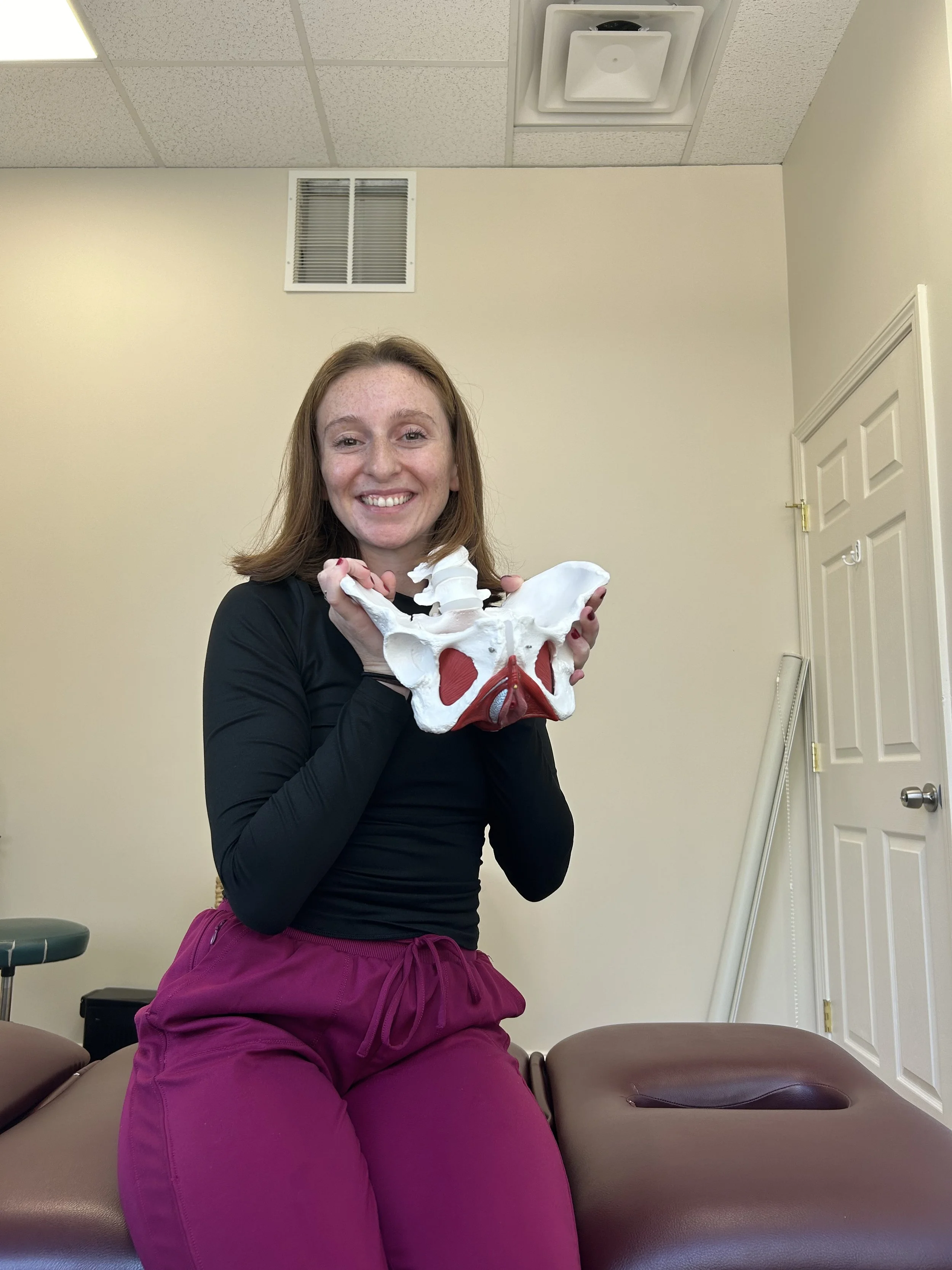 A woman smiling and holding a pelvis anatomical model in a medical office.