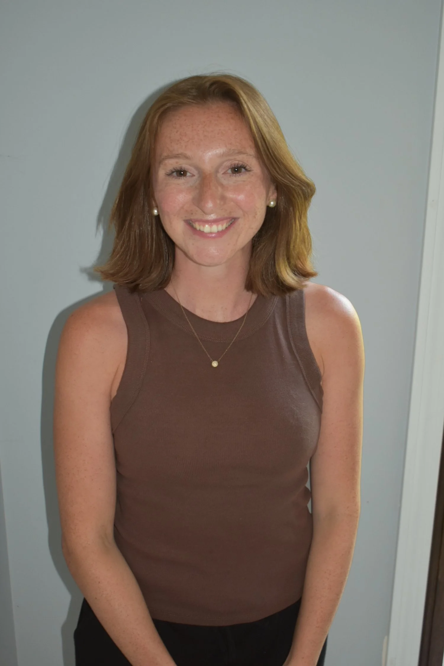 A young woman with shoulder-length red hair, freckles, and a big smile, wearing a sleeveless brown top and pearl earrings, standing against a plain wall.