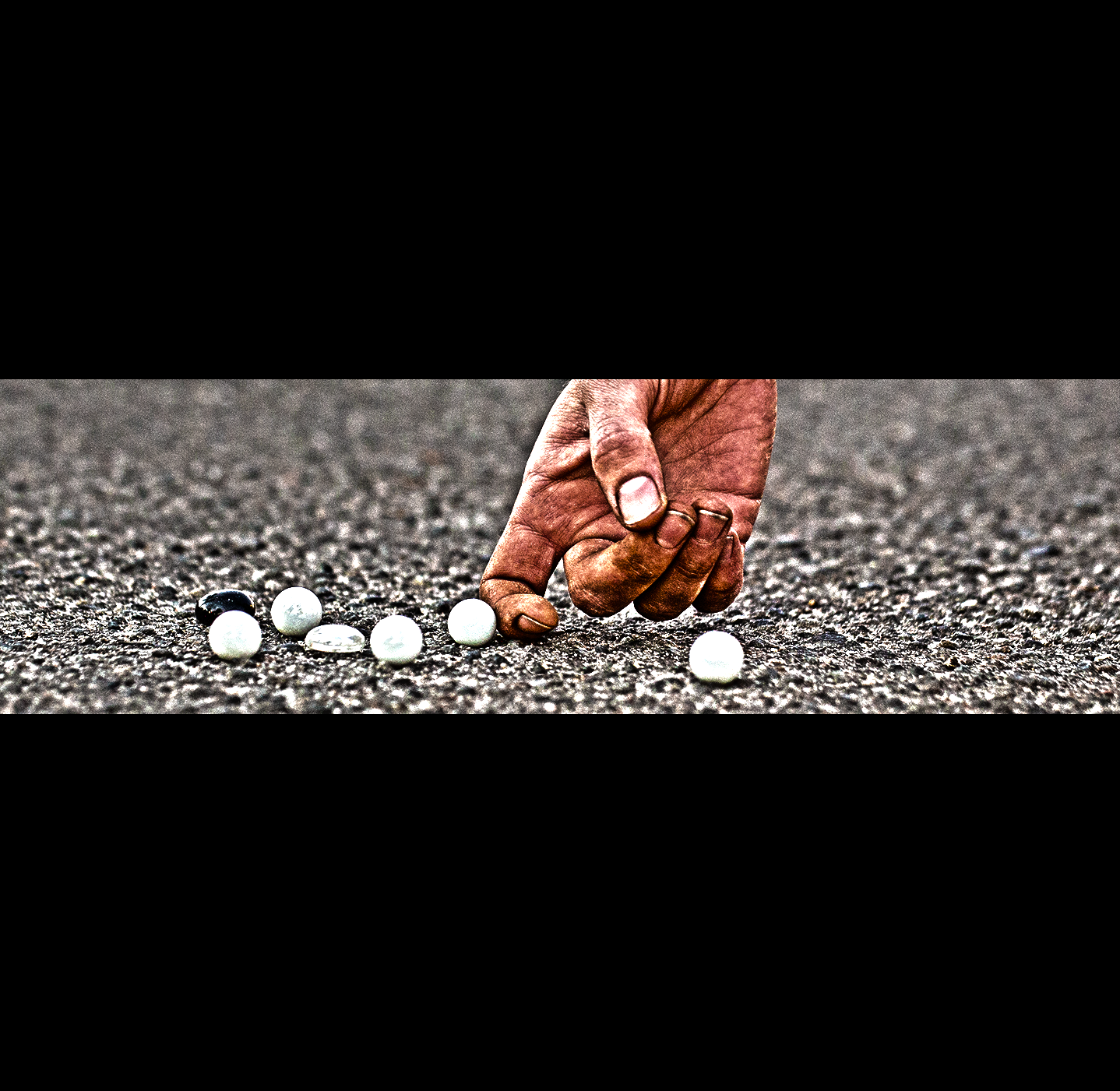 A Dirty hand shown gently nudging marbles into place on a rough asphalt road surface, close-up in daylight.