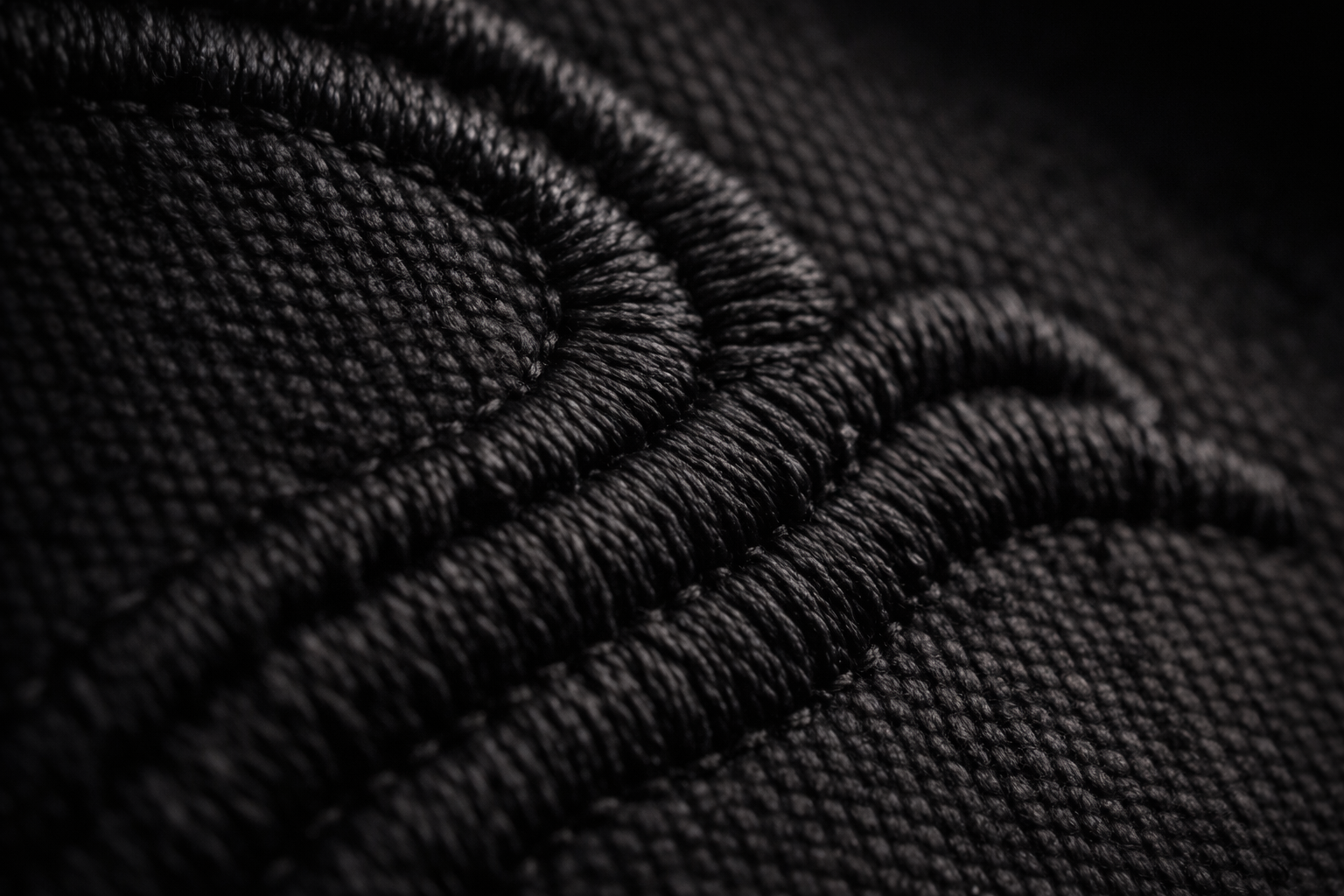 Close-up macro photograph of black embroidery stitched into heavy black cotton fabric, showing raised thread texture and dense weave detail.
