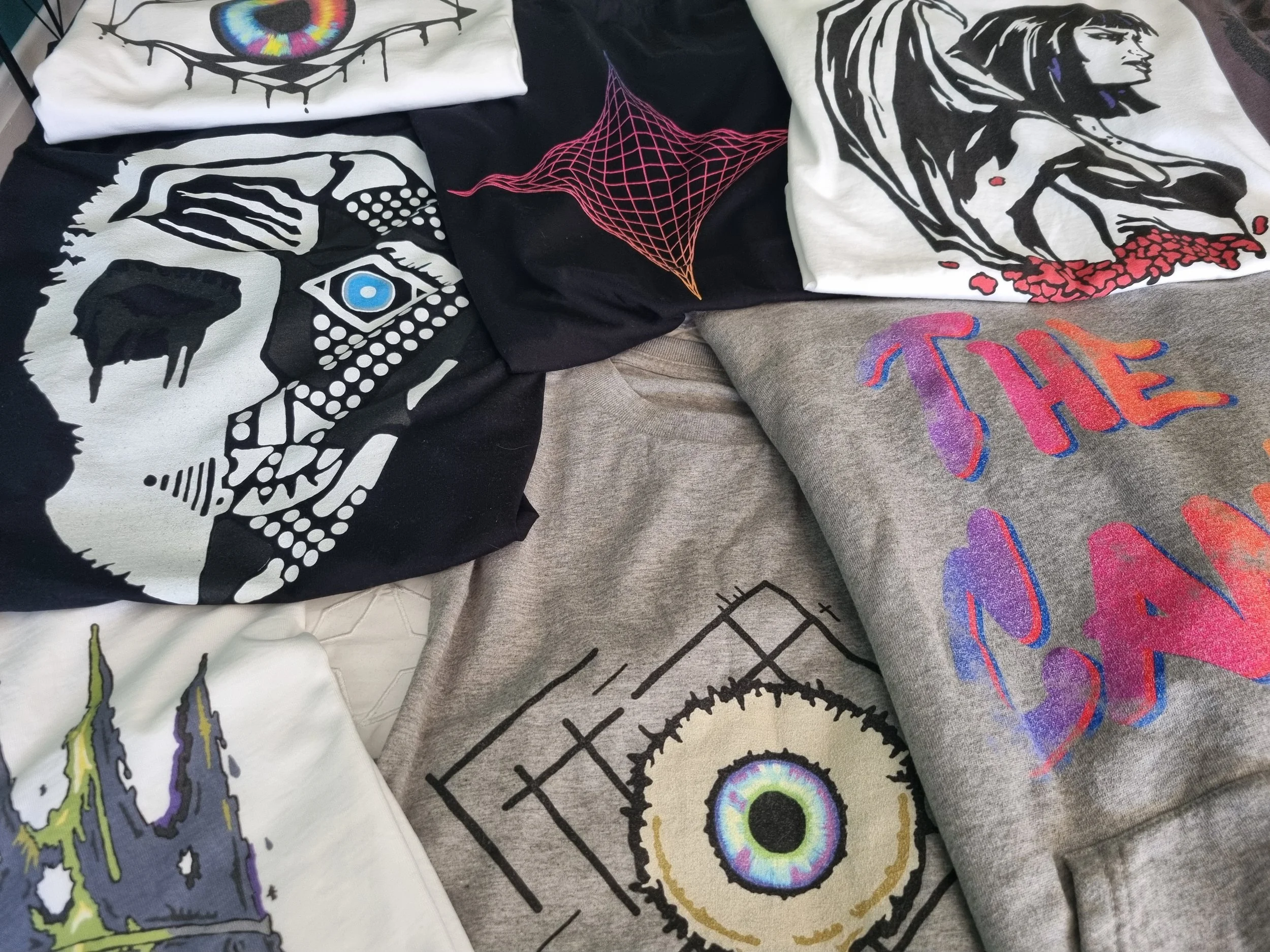 Close-up arrangement of Mongrel Logic organic cotton garments showing the Tenet 2 face, Geometric Eye, The Canis logo and other original graphic designs.