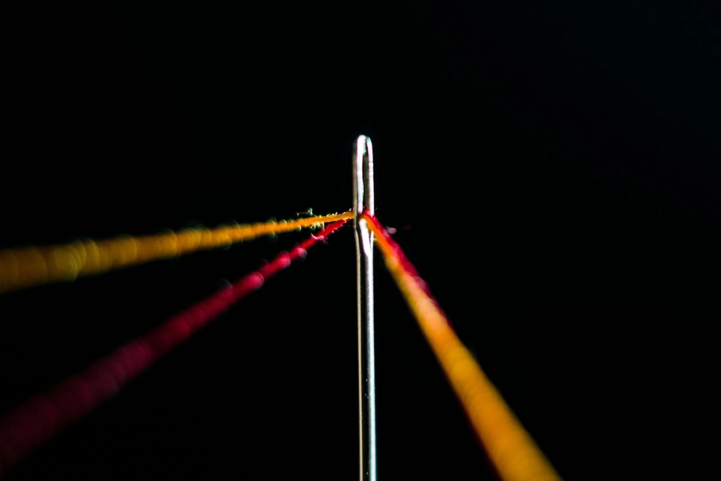Close-up macro photograph of a sewing needle threaded with red and yellow threads under tension against a black background