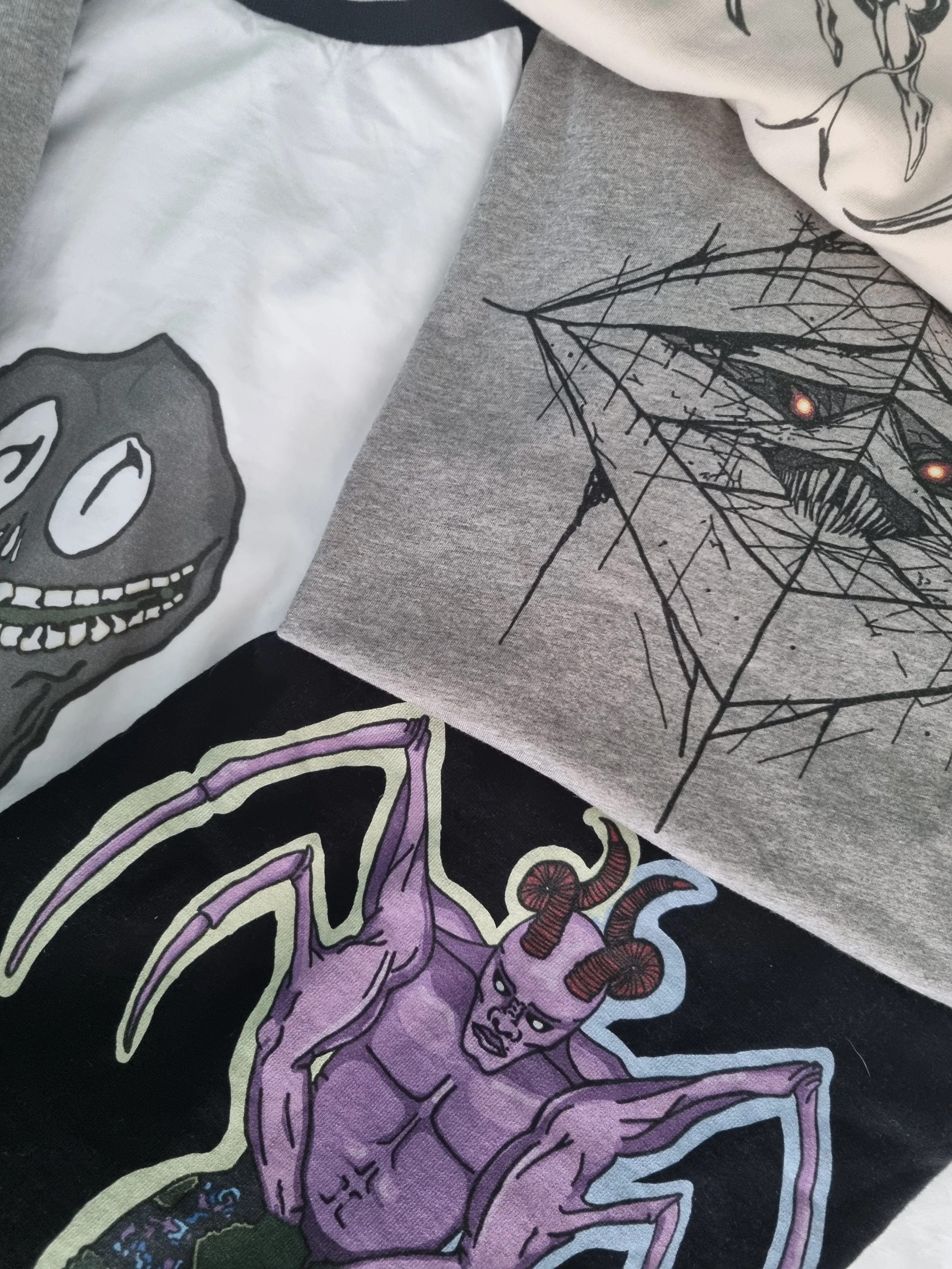 Close-up arrangement of Mongrel Logic organic cotton T-shirts and sweatshirts featuring Sun Eater, Raptor and other original dark fantasy designs.