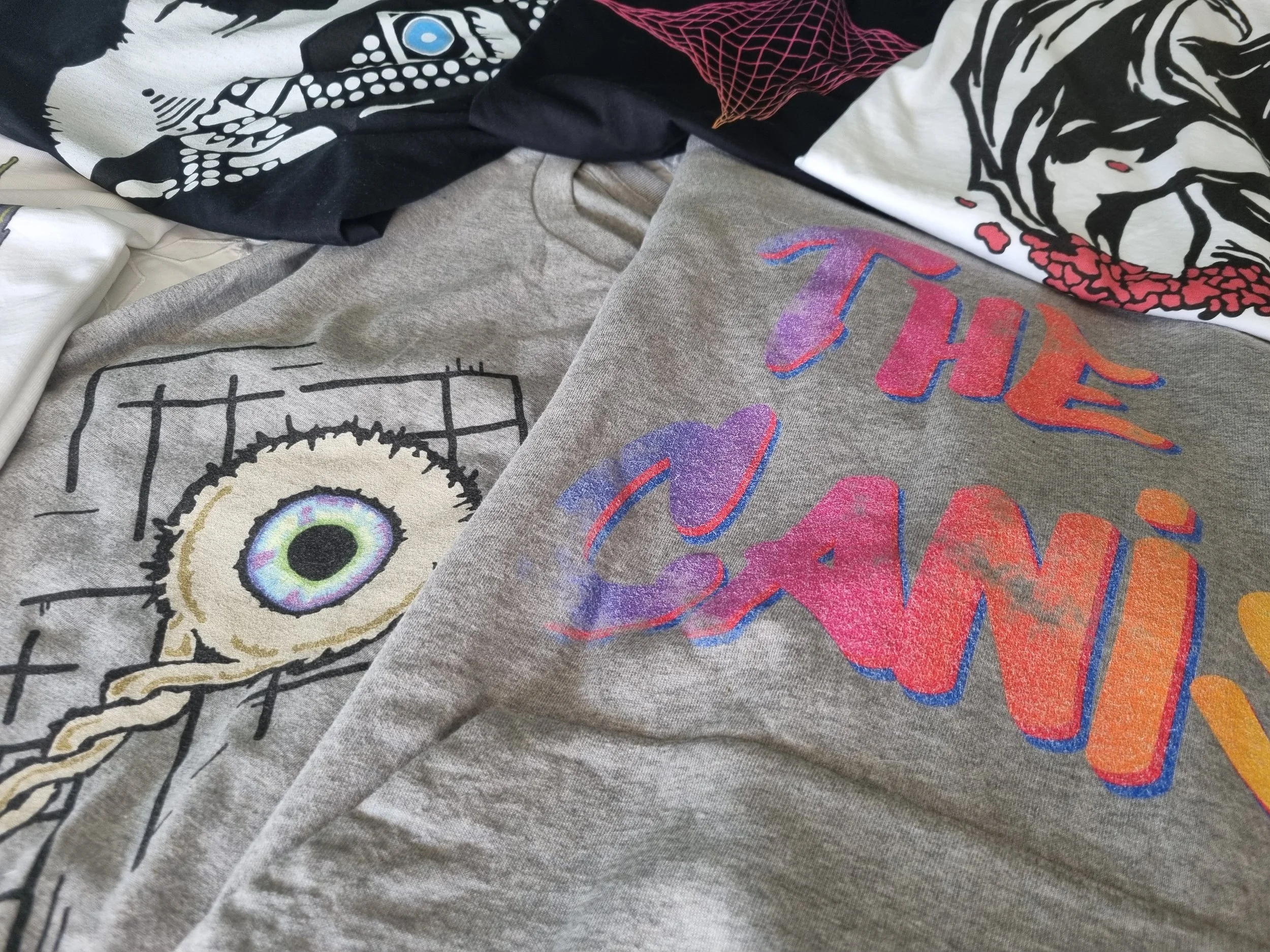 Close-up of the grey The Canis organic cotton hoodie beside black and white Mongrel Logic graphic garments featuring original artwork from The Canis.