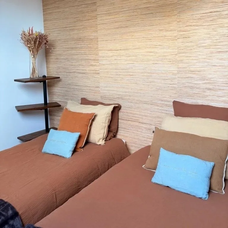 Chambre avec deux lits, chacun avec des draps marrons, des oreillers beige, marron et bleu, un tapis blanc sous les lits, sur le mur de gauche un rouleau blanc, sur le mur du fond une étagère noire avec un vase de fleurs séchées.