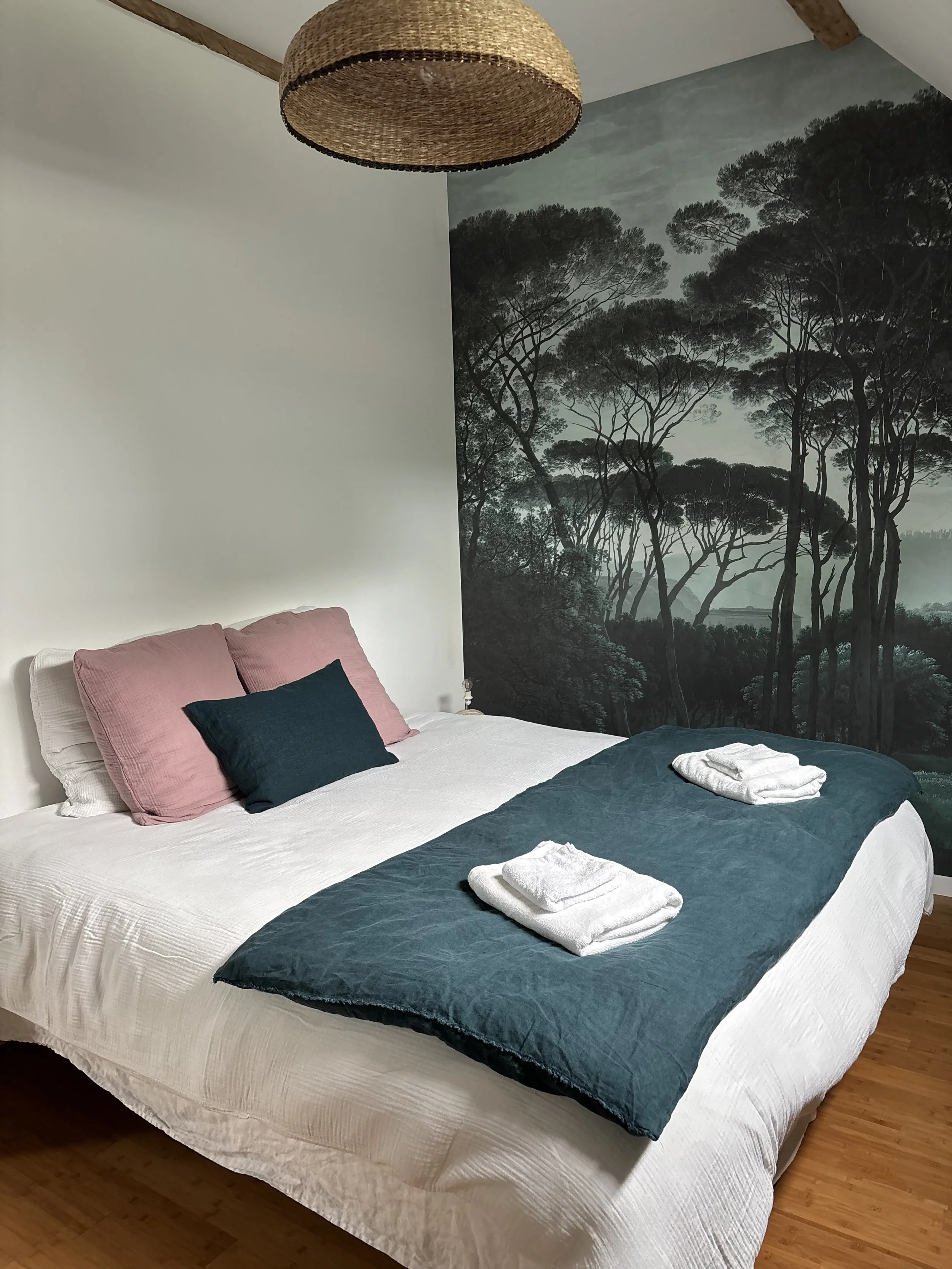 Lit double avec oreillers roses et un oreiller bleu foncé, couverture bleue avec serviettes blanches pliées sur le lit, mur avec murale de forêt de conifères.
