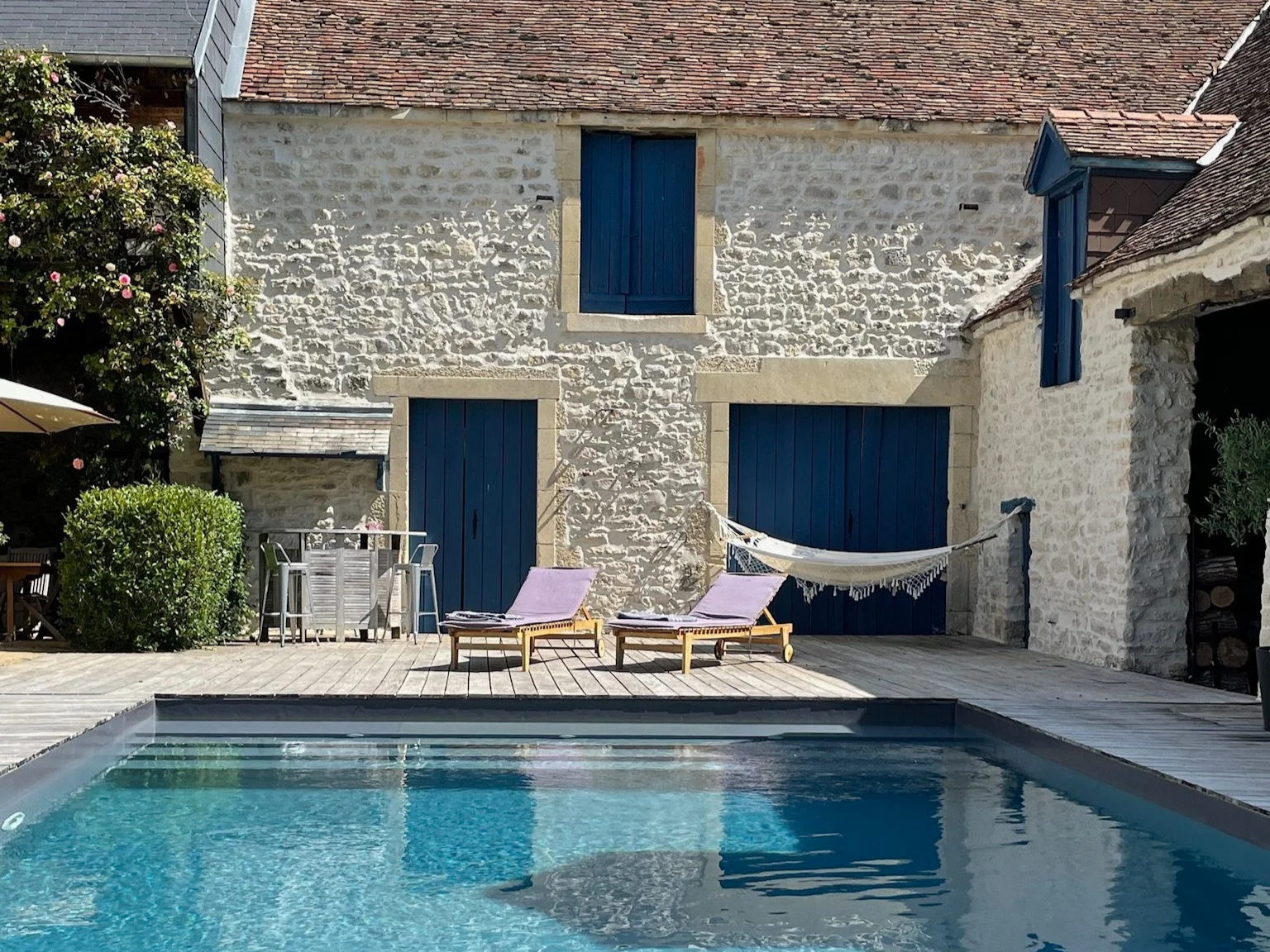 Terrasse extérieure avec une table en bois, six chaises en bois, un canapé blanc, un cadre avec une photo de palmier, et des voûtes en bois avec des murs en pierre.
