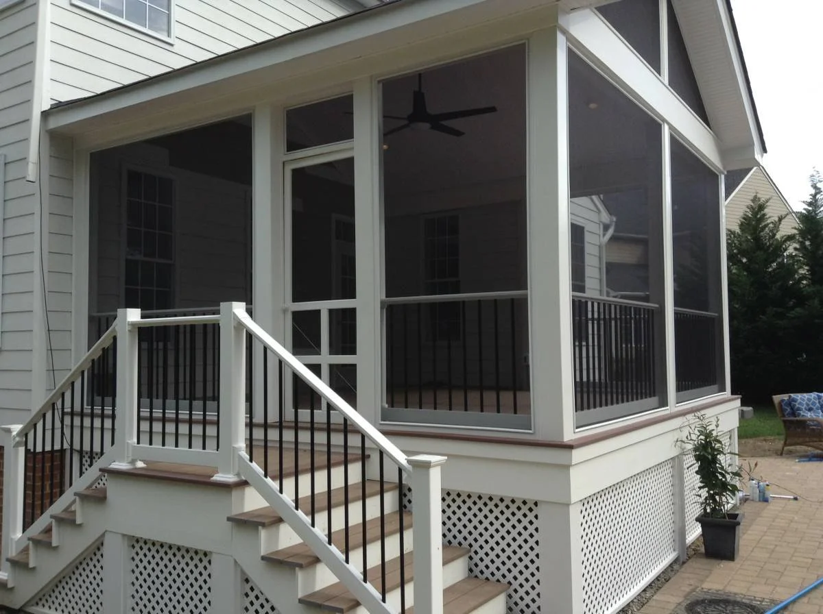 A screened-in porch with white framing, black railing, and wooden steps leading up to it, attached to a house with white siding.