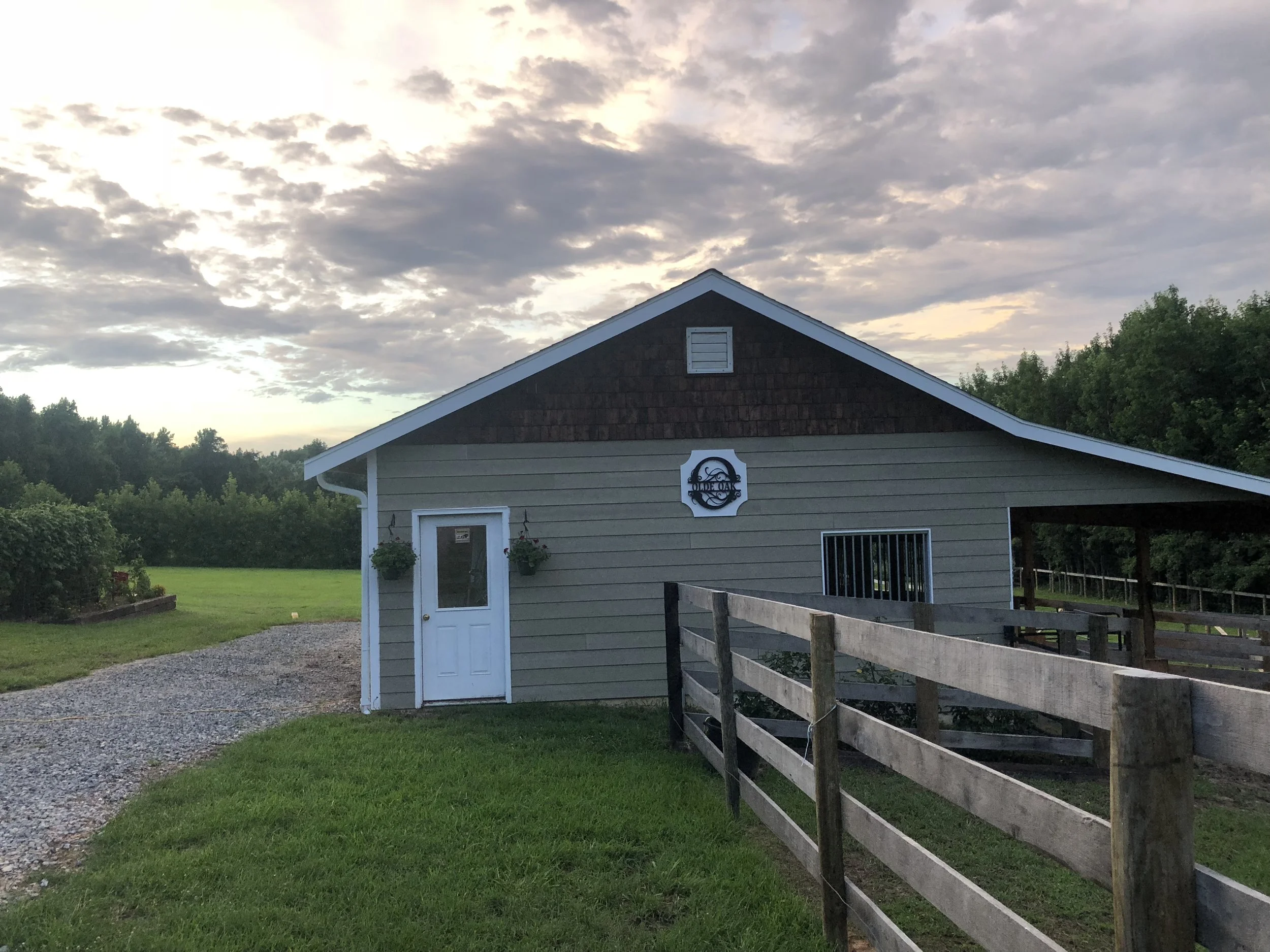 A small countryside barn with beige siding, white trim, dark shingle gable, and white door with hanging potted plants, under a partly cloudy sky at sunset, surrounded by green grass, trees, and a gravel driveway.