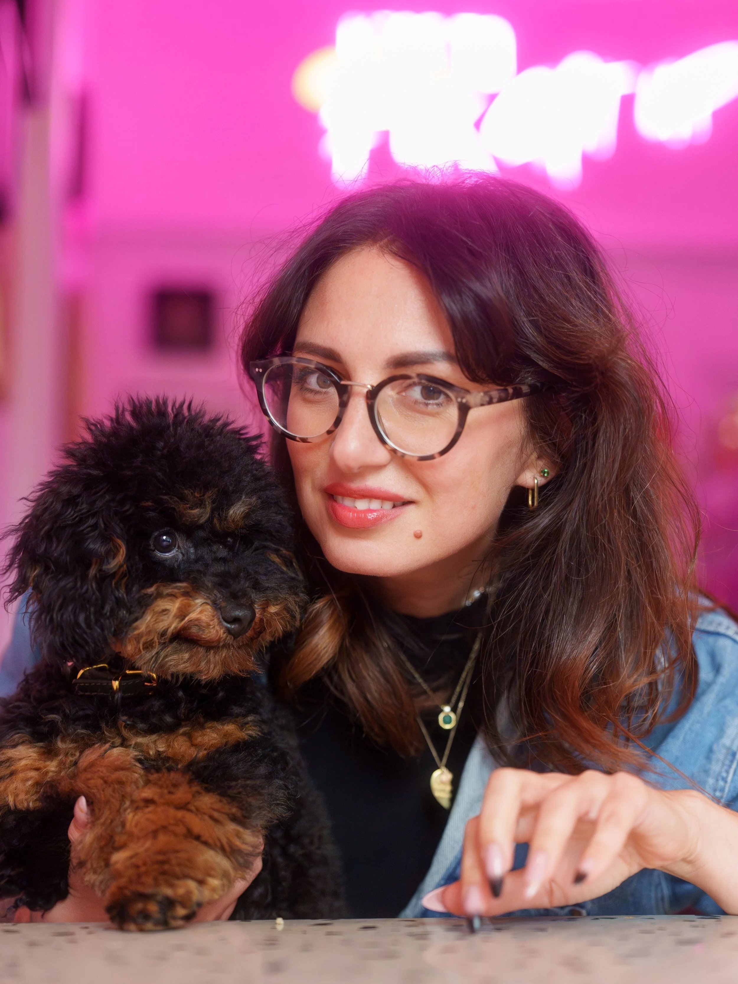 A woman with glasses and long brown hair is holding a small black and brown puppy, standing at a table with a pink neon sign in the background.