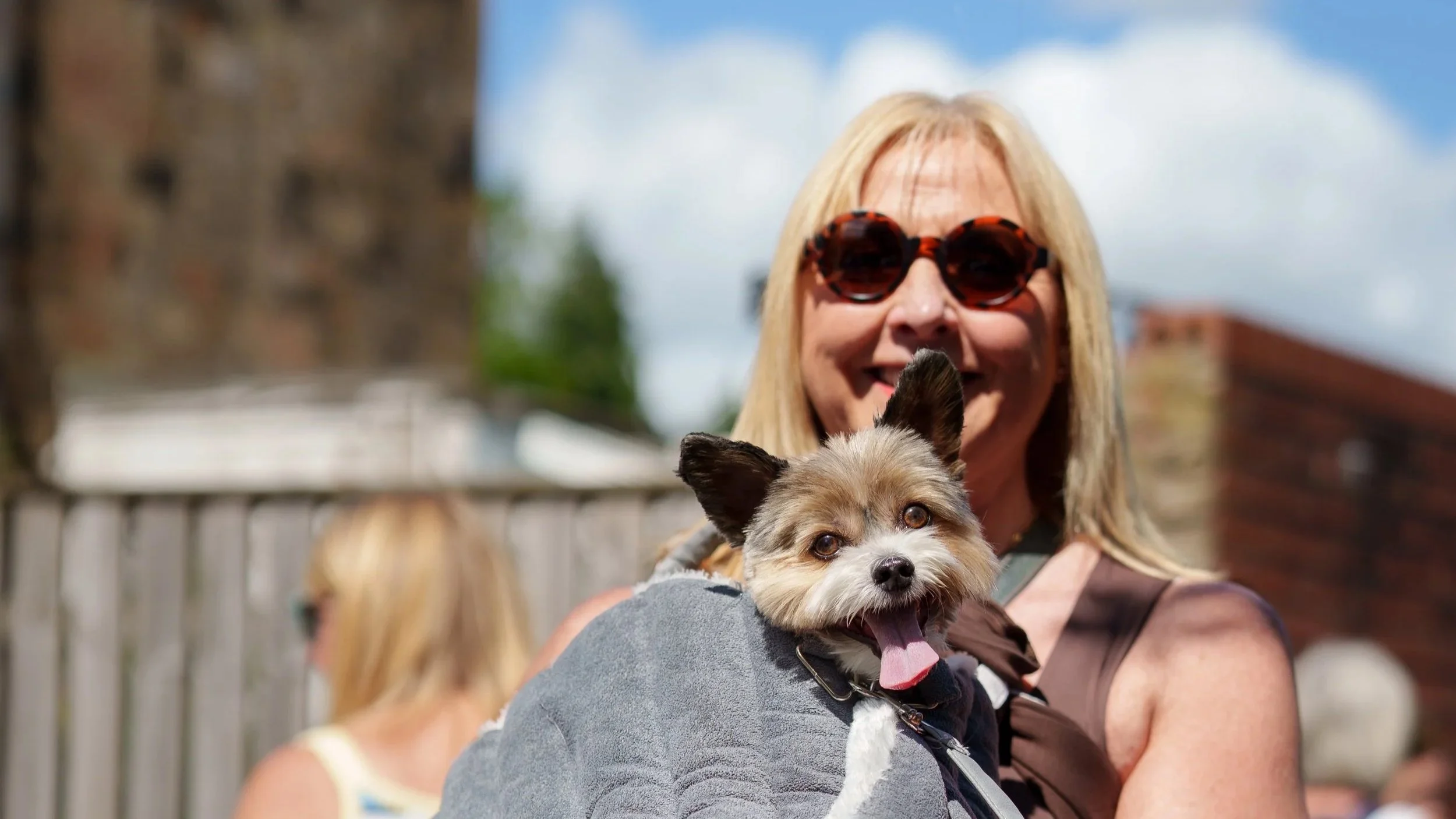 A woman with blonde hair wearing tortoise shell sunglasses is smiling and holding a small dog, which appears to be a Yorkshire Terrier mix with tan and white fur, in an outdoor setting with a cloudy sky.