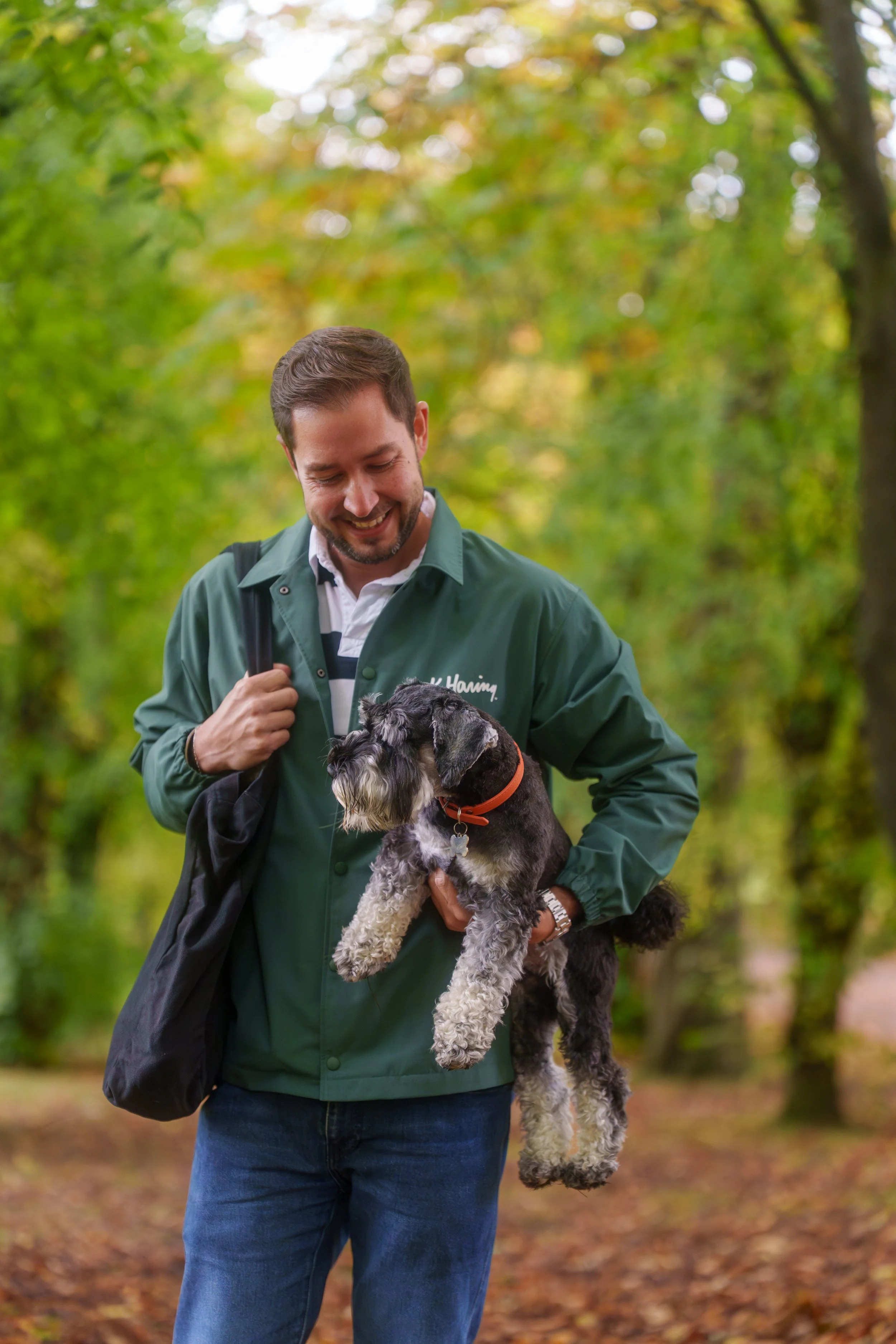 A man smiling and holding a black and gray dog while walking in a park with autumn foliage.