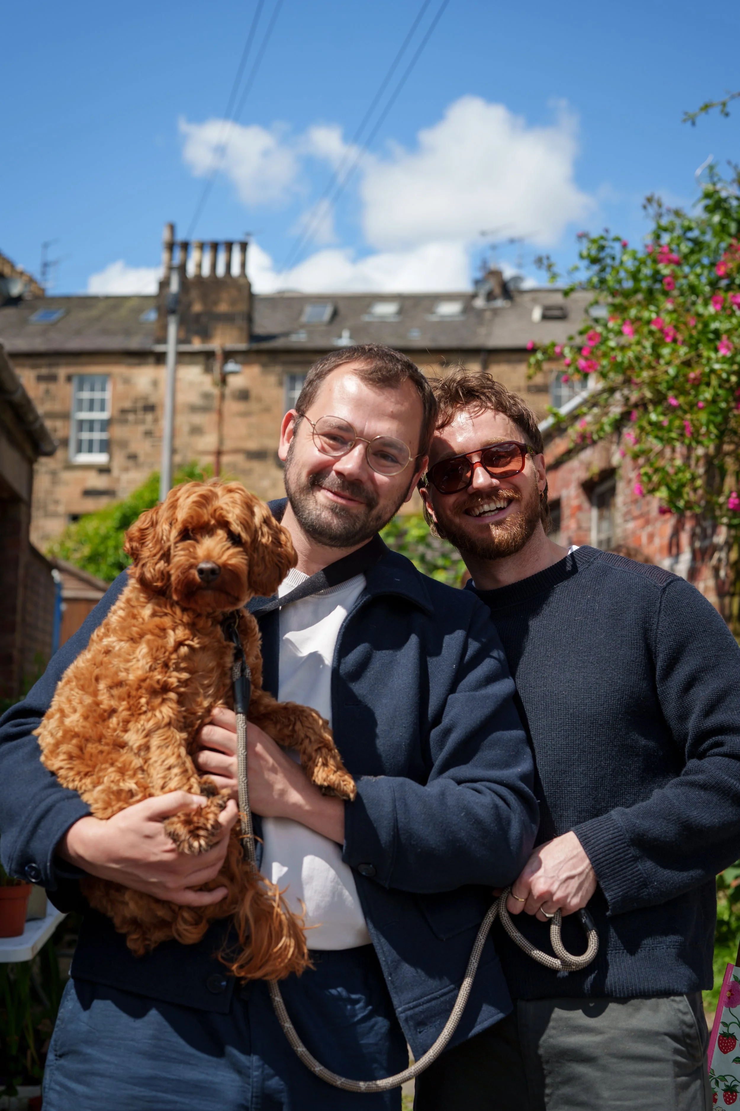 Two men smile and pose together, one holding a small brown curly-haired dog. They are outside in front of a brick building with a cloudy blue sky above and pink flowering bushes on the side.