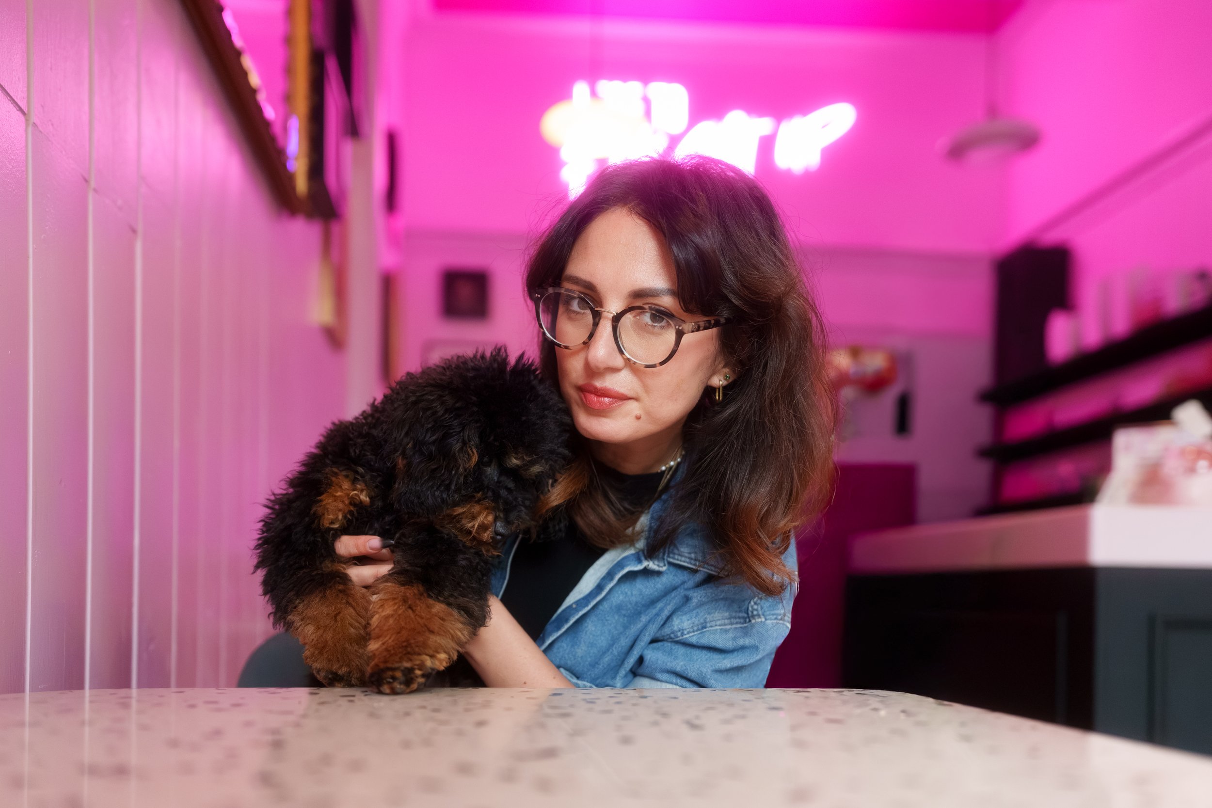 A woman with glasses and dark wavy hair holding a small black and brown puppy in a neon-lit room