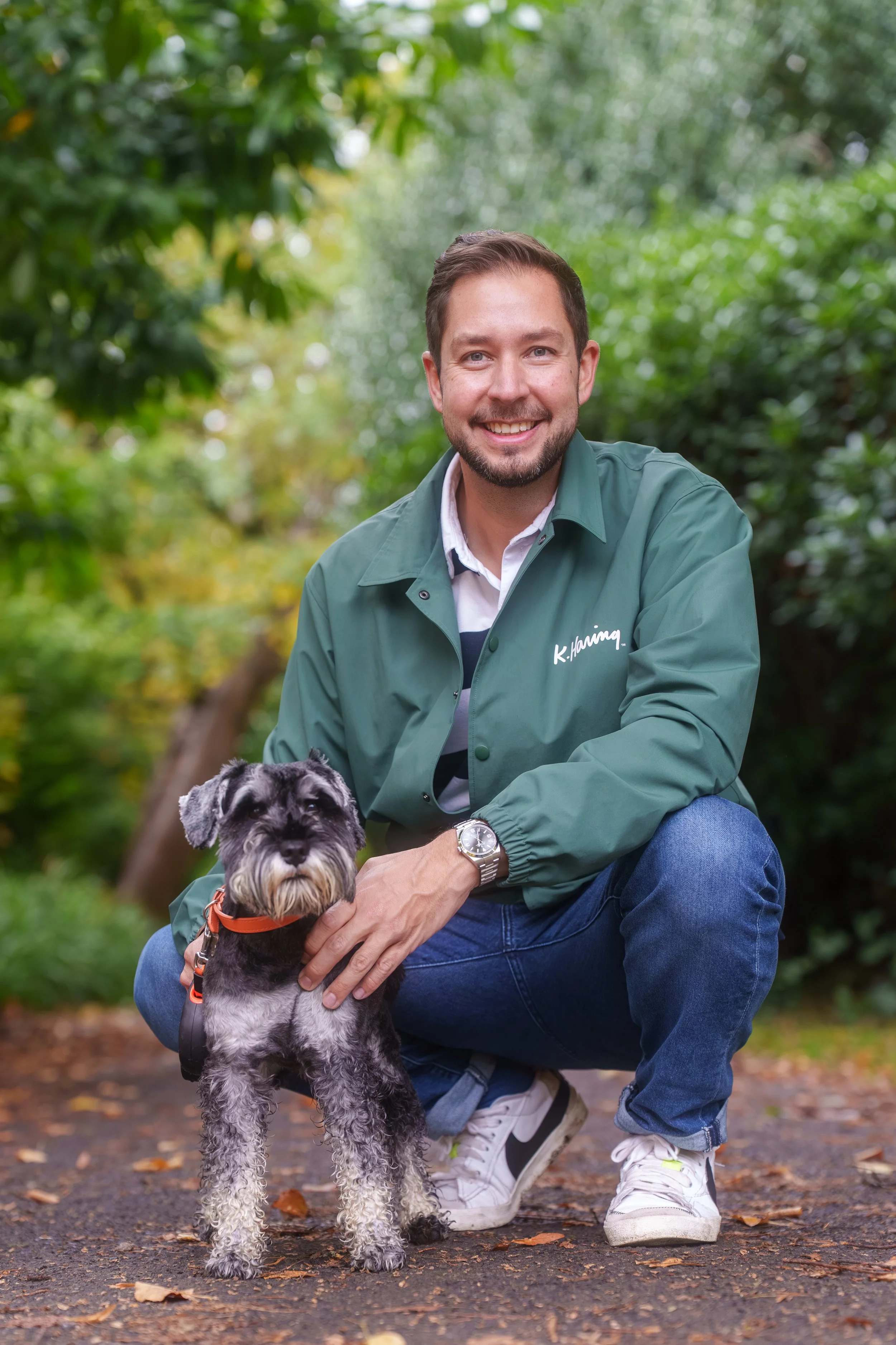 A man with brown hair and beard smiling, crouching outdoors with a Schnauzer dog, surrounded by green foliage.