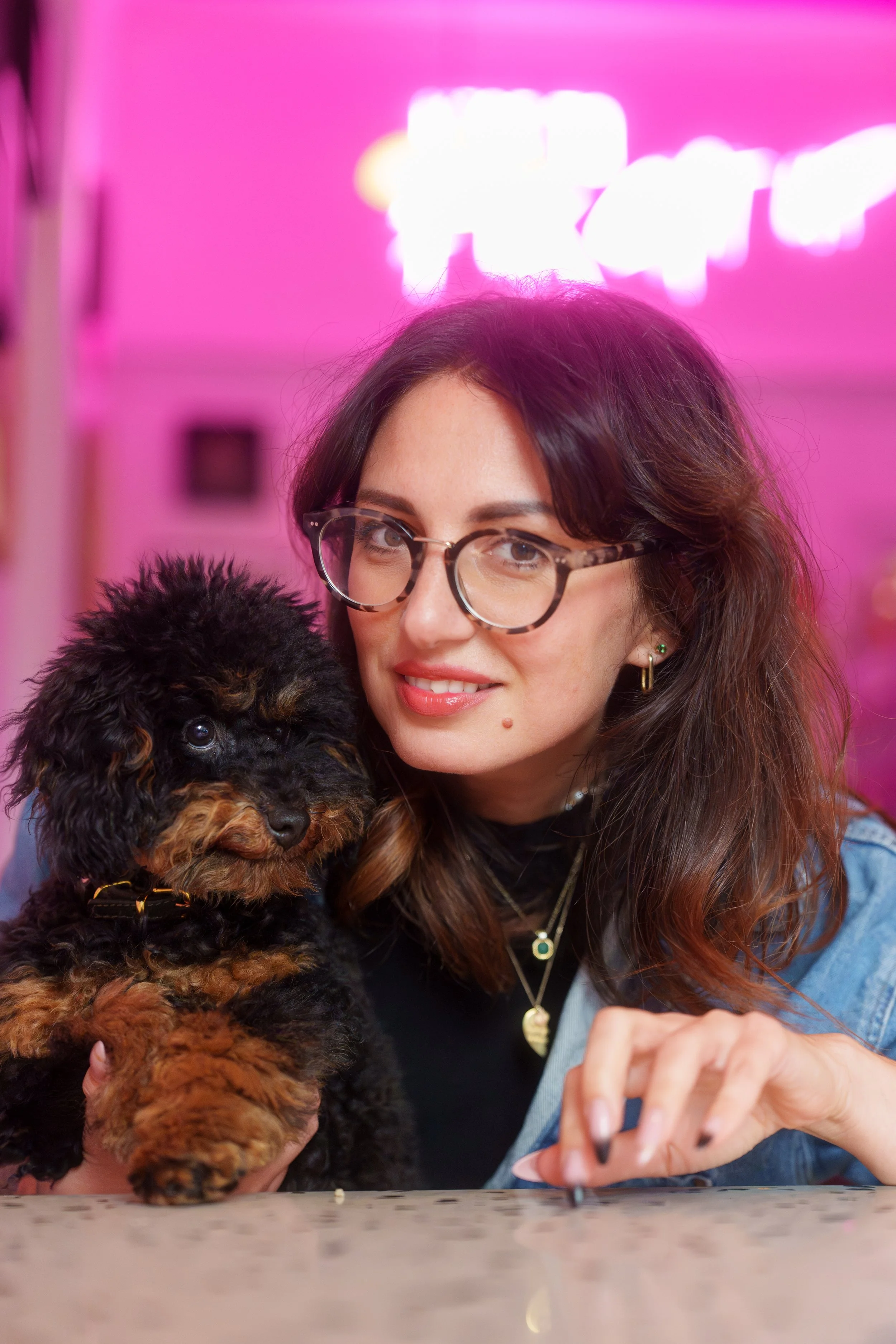 A woman with glasses and wavy brown hair holding a small black and brown fluffy puppy in a brightly lit pink room.
