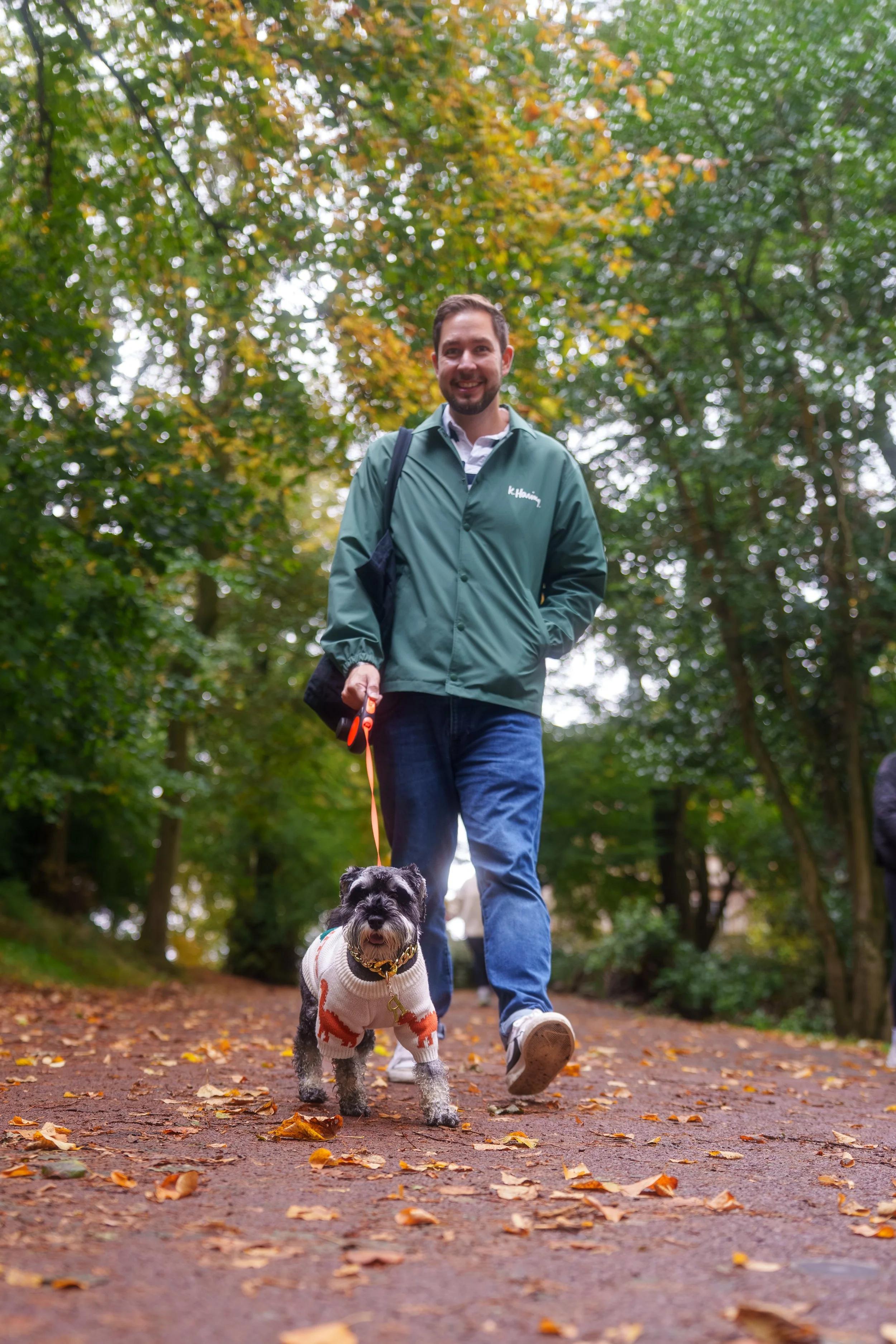 A man walking a small dog on a path through a park with trees and autumn leaves.