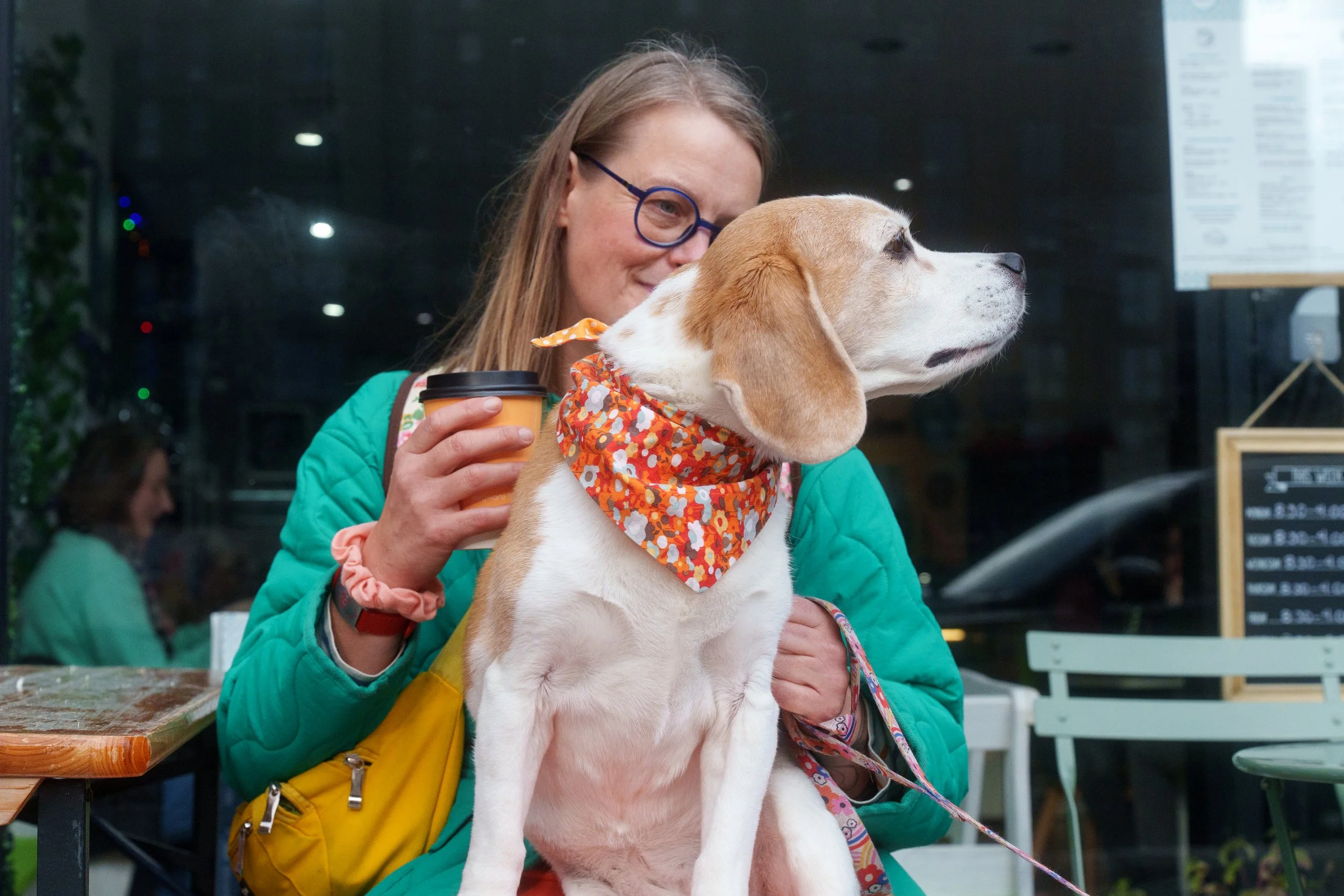 A woman in glasses and a green jacket holding a coffee cup with a beagle dog in front of her. The dog is wearing an orange floral bandana and looking to the right. They are sitting outside a cafe at night.