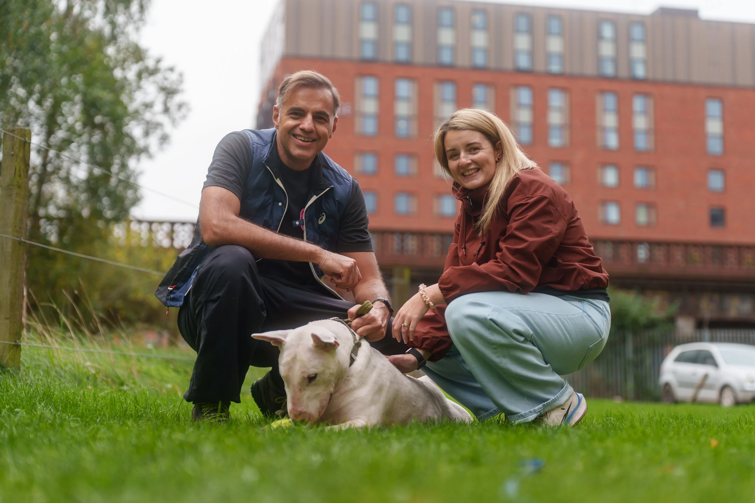 Two people, one man and one woman, crouching on green grass outdoors, smiling at the camera, with a white puppy lying in front of them on the grass, buildings and a car in the background.