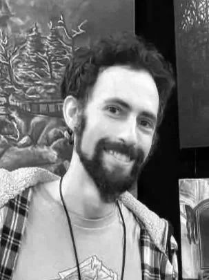 A young man with dark curly hair and a beard smiling in front of a background with trees and mountain scenes.